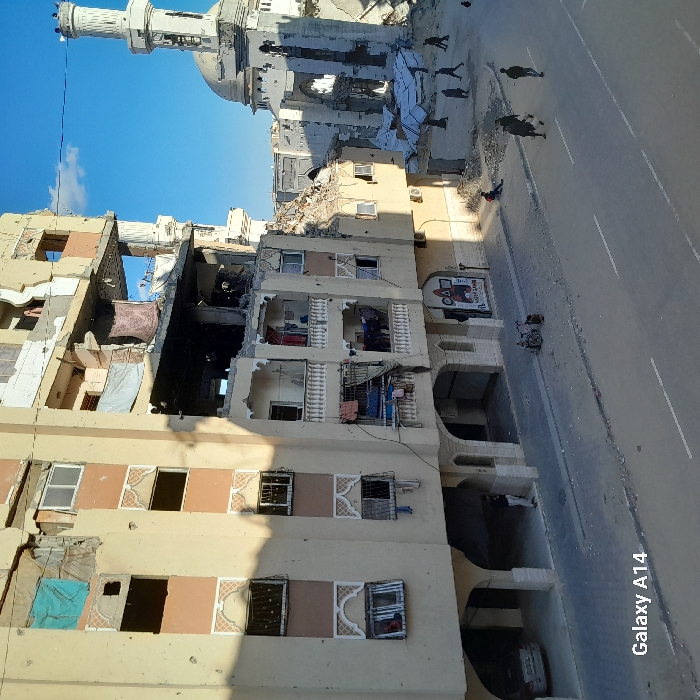 The image depicts a devastated urban street scene in Khan Yunis, Palestinian Territory, under a clear blue sky. On the left, a multi-story building, at least six floors high, exhibits extensive structural damage. Its beige facade, featuring brown decorative panels and arched window frames, is pockmarked with bullet holes and shrapnel marks. Large sections of its walls are missing, exposing internal rooms, while many windows are shattered or completely absent. Balconies on the middle floors are visible, with some showing laundry hanging, including blue and pink fabrics. The upper floors are particularly ravaged, with debris and fabric visible through gaping holes. At ground level, the building features arched open bays; a partially visible white vehicle is parked in one, and a person in a white garment stands under another arch. A faded sign for "MAZIN GAMES" is discernible on the ground floor facade.
To the right of the damaged building stands a heavily ruined mosque. Its distinct minaret is partially intact, but the dome and significant portions of the main structure are collapsed or missing, exposing internal frameworks. A large pile of concrete slabs and other debris lies scattered at the mosque's base.
The street in the foreground is paved, with visible but worn white lane markings. The adjacent sidewalks are also damaged and strewn with some smaller debris, particularly near the mosque. Approximately 10-12 individuals are present in the scene. Several figures are walking on the street and sidewalks, generally moving away from the viewer or across the street. One person is seated on the ground amidst the debris near the mosque. A bicycle, possibly with a child seat, is leaning against the curb near the multi-story building. Shadows cast by the buildings suggest it is daytime.