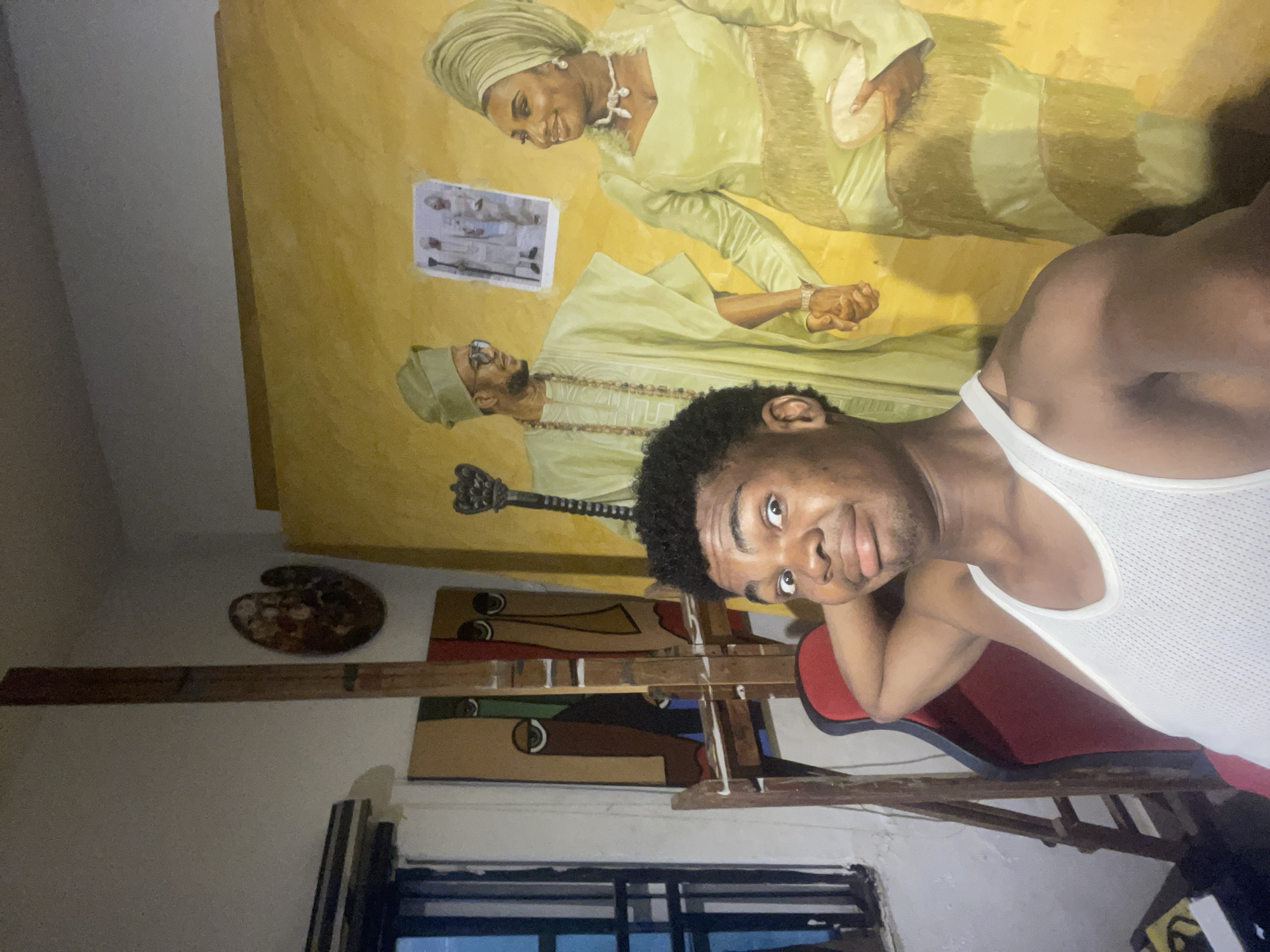 This is a medium shot, selfie-style photograph taken in an artist's studio, likely in Ado, Nigeria. A young Black man, wearing a white tank top, is in the foreground, looking at the camera with a playful expression and his arm behind his head. He is sitting in a red and black racing-style chair. Behind him, an easel holds two abstract paintings of faces with distinct eye designs. To the right, a large, unfinished oil painting on canvas depicts a Black couple dressed in traditional Nigerian attire. The man in the painting wears a light green agbada and cap, while the woman wears a matching light green iro and gele. They are holding hands, and a small framed photograph of a similar couple is affixed to the canvas. Other art supplies, including a painter's palette, are visible on the wall in the background. The lighting suggests an indoor setting, possibly with natural light coming from a window on the left side of the frame.