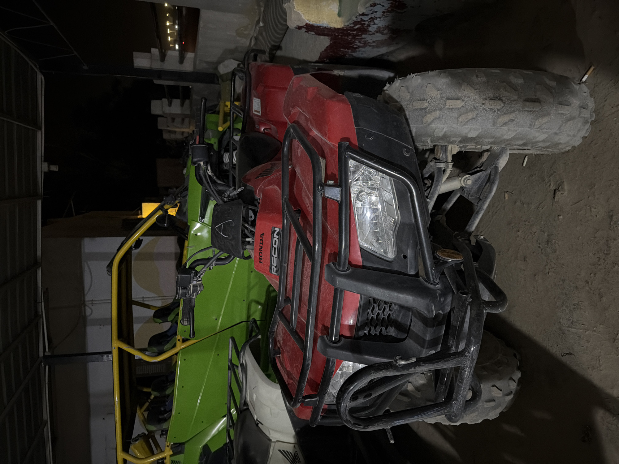 A red Honda Recon ATV is parked in the foreground, facing forward. It has a black roll cage and black handlebars. The ATV is dirty and appears to be used. Behind it is a green UTV with a yellow roll cage, also facing forward. The UTV has multiple seats. Both vehicles are parked indoors, possibly in a garage or storage unit. The background is dark and appears to be a rough, concrete floor. The overall impression is one of a storage area for recreational vehicles. The location is identified as Sharm El Sheikh, Egypt, though there are no specific visual cues to confirm this. The scene is lit by artificial lights, creating shadows and highlighting the textures of the vehicles and the environment.