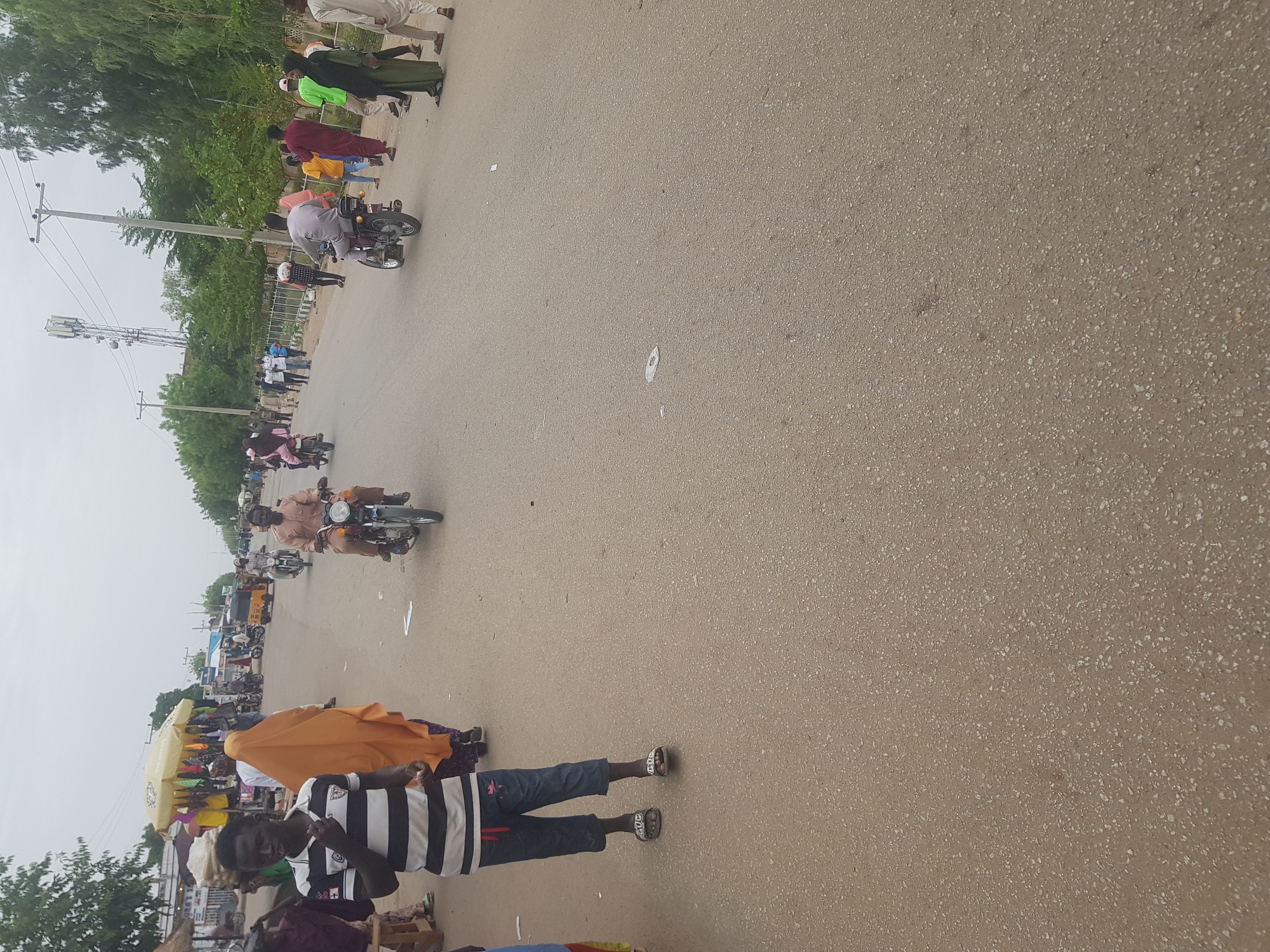 The image captures a street scene in Dabawa, Nigeria. It appears to be a daytime shot with overcast lighting. The main subject is a paved road, partially filled with people and motorcycles. Some individuals are walking along the edge of the road, while others are riding motorcycles. A small market area is visible in the background. The presence of power lines and utility poles are also noticeable along the roadside.