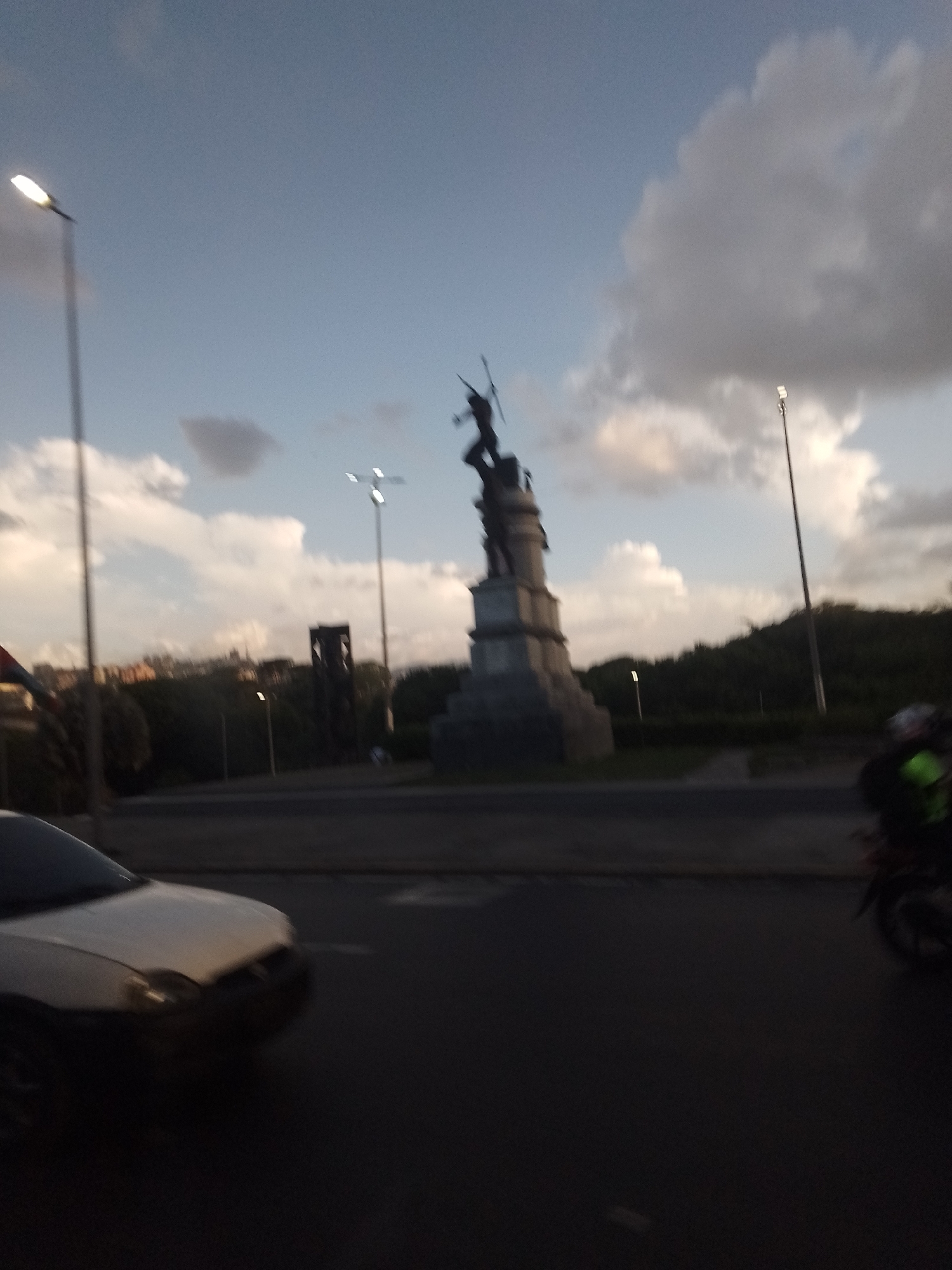 The image captures an urban scene in Caracas, Venezuela, during what appears to be dusk or early evening. The sky is a pale blue with scattered, large white and gray clouds, and some streetlights are already illuminated.
In the center of the frame stands a prominent dark-colored statue on a multi-tiered stone pedestal. The statue depicts a figure, possibly a male, holding a long spear or staff, with an arm raised. This monument is situated within a circular road or roundabout.
Around the monument, several tall streetlights are visible, with their bright lamps glowing against the dimming sky. To the left, a light-colored car is partially visible in the bottom left foreground, appearing to be in motion, indicated by motion blur. On the far right, part of a motorcycle or scooter, along with its rider, who seems to be wearing a neon green or yellow vest, is also blurred, suggesting movement.
The background features a line of dark green trees and dense vegetation, creating a silhouette against the horizon. Behind the main monument and further back, another distinct dark structure, possibly a part of a building or an abstract monument, stands out against the sky. The overall scene suggests a busy city intersection centered around a historical or monumental feature. No visible text is present in the image.