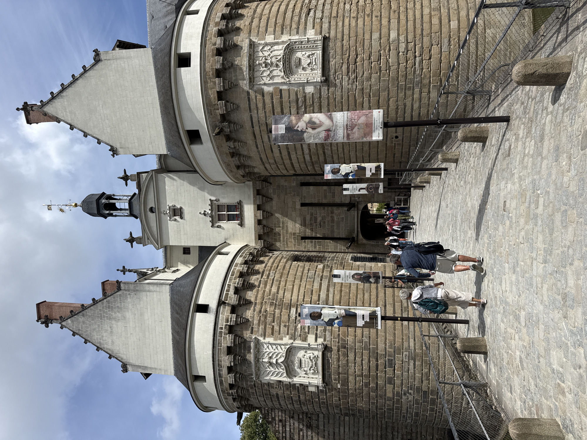 This image shows the entrance to the Château des Ducs de Bretagne in Nantes, France, on a bright, cloudy day. A cobblestone bridge leads to the castle's fortified entrance, flanked by two large, round stone towers. The towers have ornate stone carvings above arched doorways, and banners advertising an exhibition are displayed on poles along the bridge.
Several people are walking towards the castle entrance. In the foreground, a couple is walking away from the camera, the woman with a green backpack and the man with a dark backpack. Further ahead, a group of people are seen entering the castle. The castle itself is built of light-colored stone and features steep, pointed roofs and a central clock tower. The overall scene depicts a historical landmark bustling with visitors.