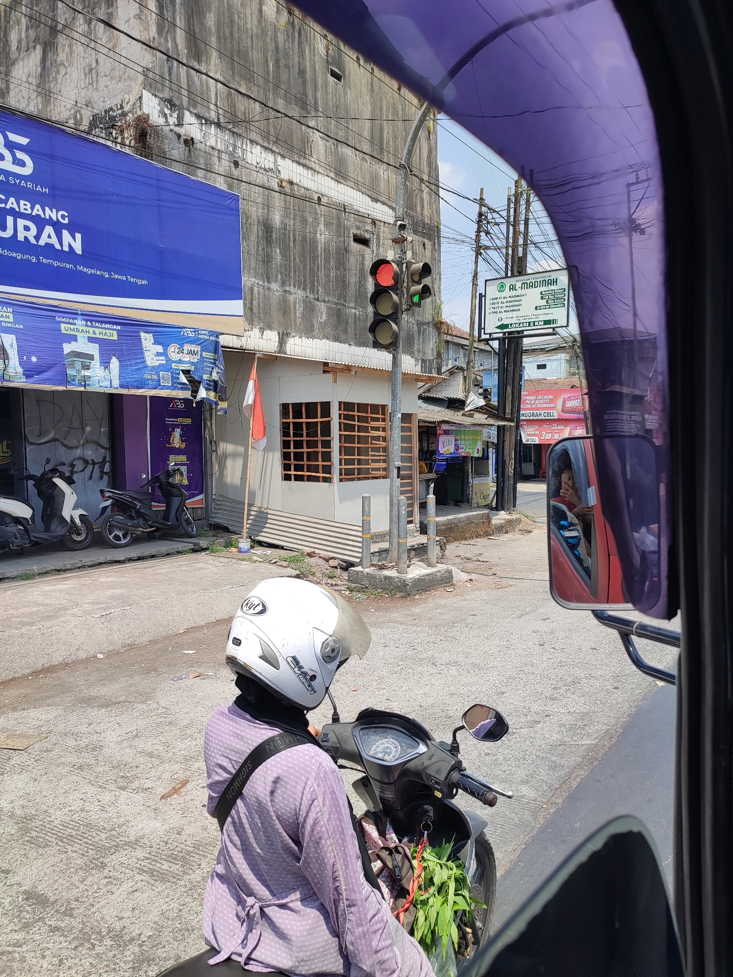 The image depicts a street scene in Tanggulrejo, Indonesia, observed from the perspective of someone inside a vehicle. In the foreground, a person wearing a white helmet and a light purple, polka-dotted long-sleeved shirt is seated on a black scooter. The scooter has a green bunch of leaves hanging from the front. To the left of the scooter, another scooter is parked.
Behind the scooters, a building with a weathered, grey concrete facade is visible. A large blue banner with white text is affixed to the building. Further back, a traffic light shows a red light and a green light simultaneously. A sign for "YAYASAN PENDIDIKAN AL-MADINAH" is mounted on a pole. In the reflection of a side-view mirror, a person can be seen with their face towards the camera. The street is paved with asphalt and appears to be an urban environment.