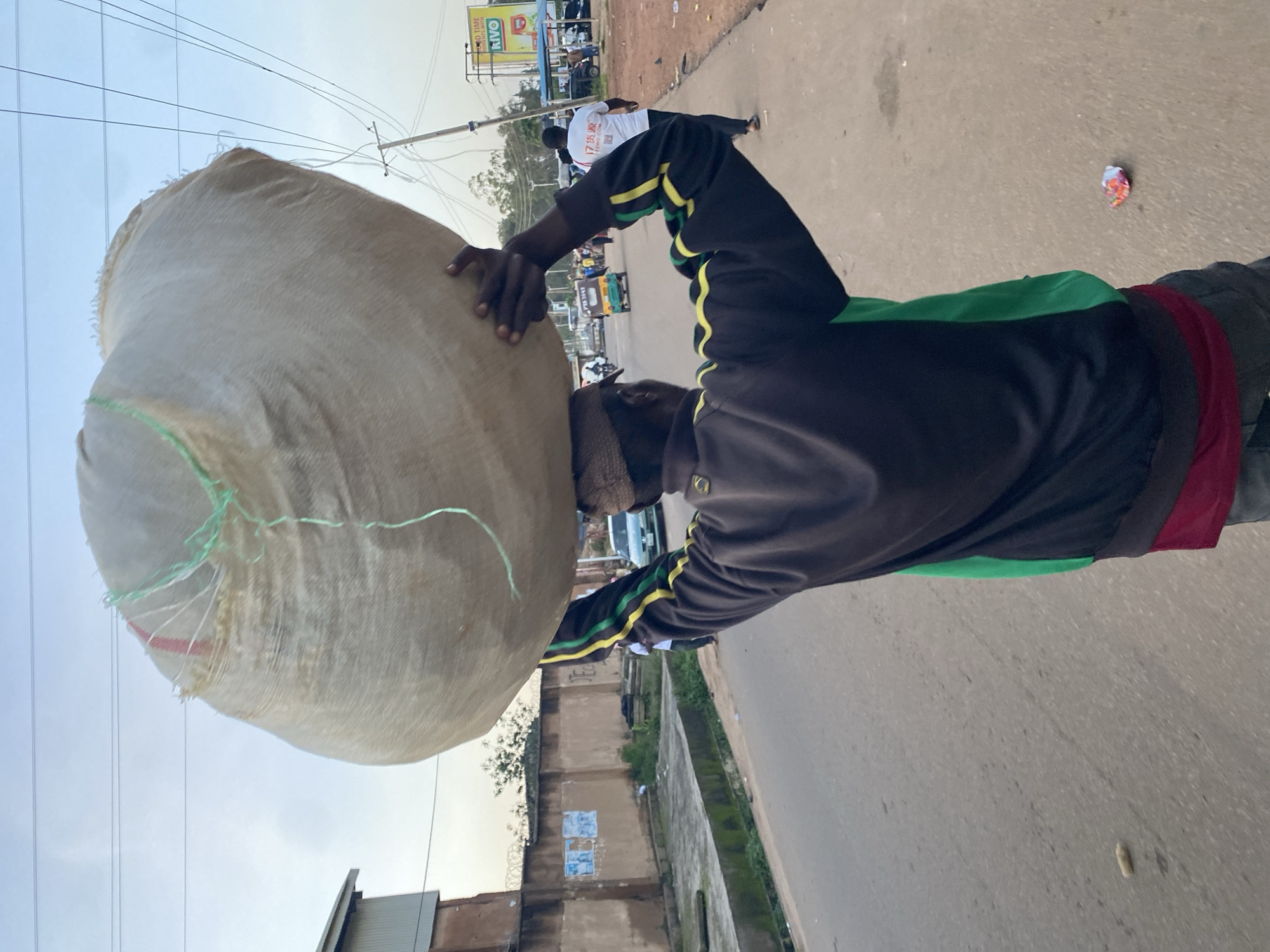 A man walks down a paved street carrying a large, light-colored sack on his head. He is wearing a dark tracksuit with green and yellow stripes on the sleeves and a red undershirt. The sack is made of woven material and tied with green string. He is walking away from the camera, towards a distant intersection with traffic, including a green auto-rickshaw. Buildings and a barbed-wire fence line the left side of the street. The sky is overcast and appears to be either early morning or late afternoon. The scene is consistent with an urban or semi-urban environment in Gura Top, Nigeria.