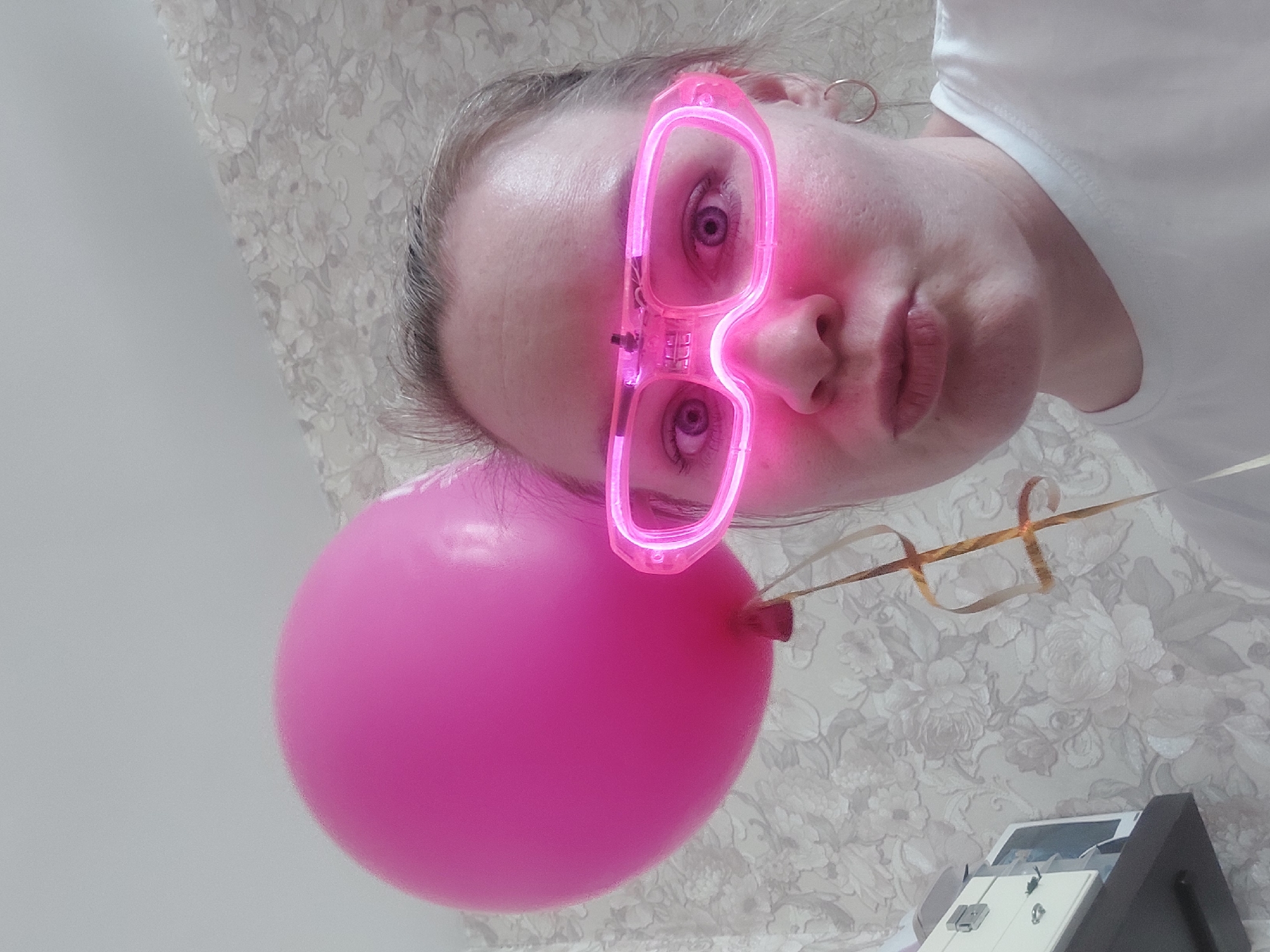 The image shows a person with light skin, wearing pink neon glasses and a white top. They are holding a pink balloon with gold ribbons. The background is a light-colored wallpaper with a floral pattern. A portion of a white and gray object is visible in the bottom right corner. The person appears to be indoors, possibly in a home. There is no visual information to suggest the location is Chaikovsky, Russia.