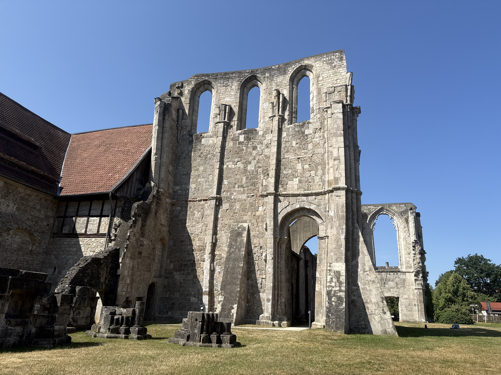 The image shows the ruins of the Walkenried Abbey, a former Cistercian monastery located in Walkenried, Germany. The abbey is characterized by its large stone structures, including a main ruin with arched windows and a partially intact roof on an adjacent building. The environment consists of a grassy field with scattered stone debris. The sky is clear and blue, indicating a sunny day. There are no people visible in the image.