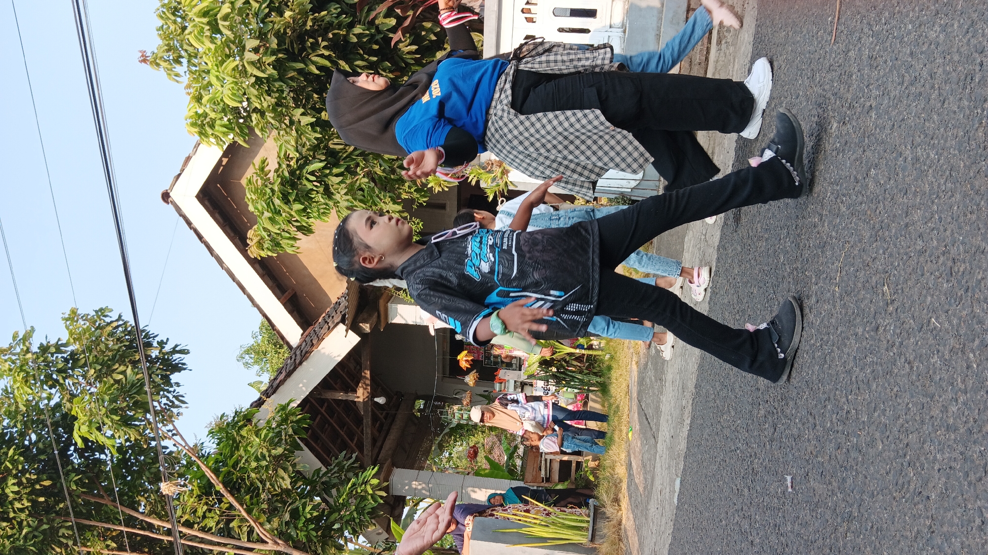 A group of people, including children and adults, are walking down a paved street in Cerme, Indonesia. The scene appears to be outdoors, with houses and trees visible in the background. The sky is clear and blue, suggesting a sunny day. The people are dressed in casual clothing, with some wearing headscarves. One young girl in the foreground is wearing a black t-shirt with a graphic design and black pants. She is walking with her head turned to the side, looking off-camera. Behind her, an adult woman wearing a blue t-shirt and black pants, with a plaid shirt tied around her waist, is also walking. Several other individuals, including children, are visible in the middle ground and background, also appearing to be walking. The overall impression is of a casual gathering or procession on a street.