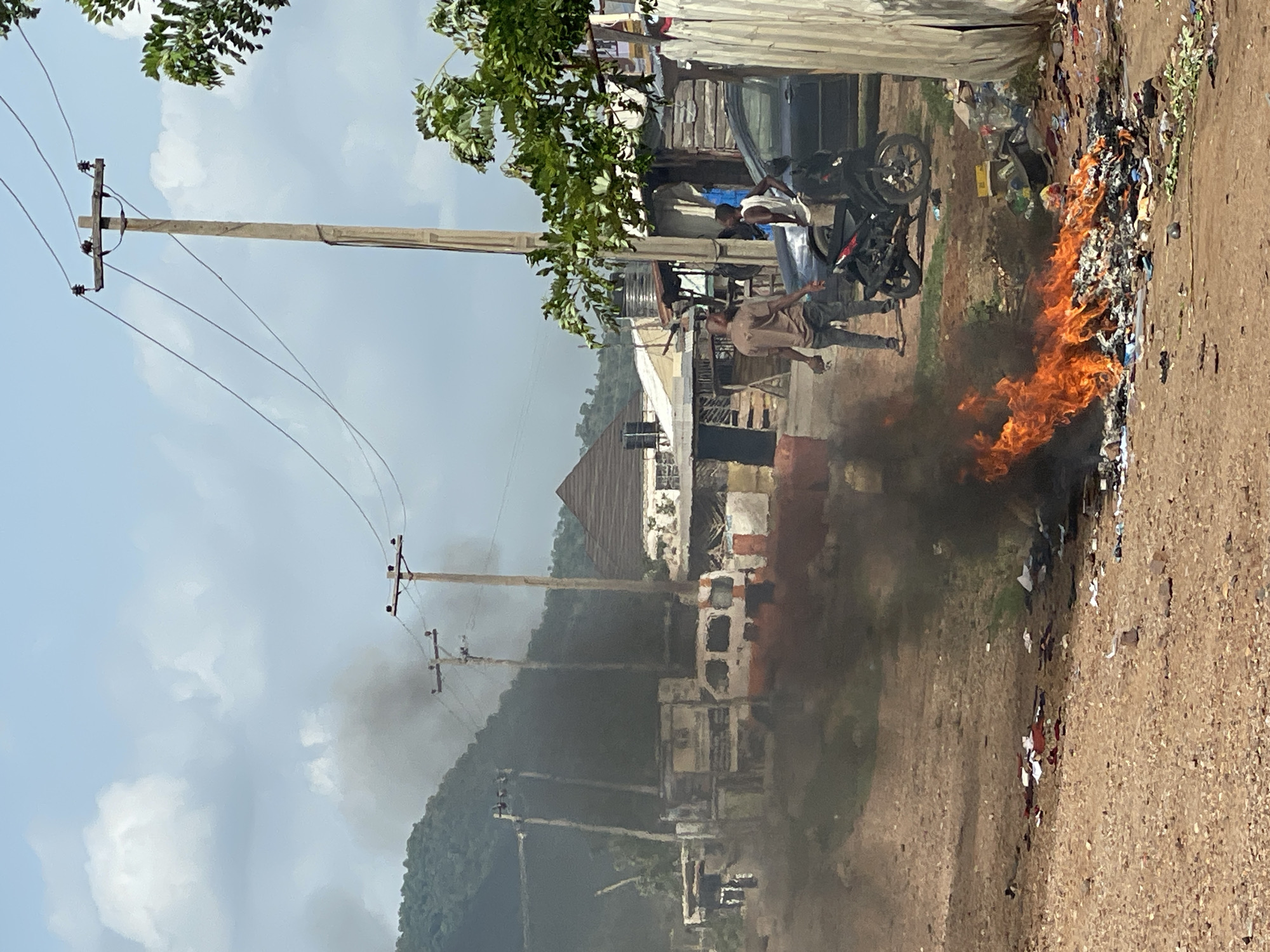 The image depicts a street scene in Lokoja, Nigeria. A fire burns in a pile of trash at the edge of a dirt road. Two men stand near a motorcycle parked next to the fire. Power lines are visible along the street. Buildings and a wooded hillside are in the background. Smoke from the fire fills the air.