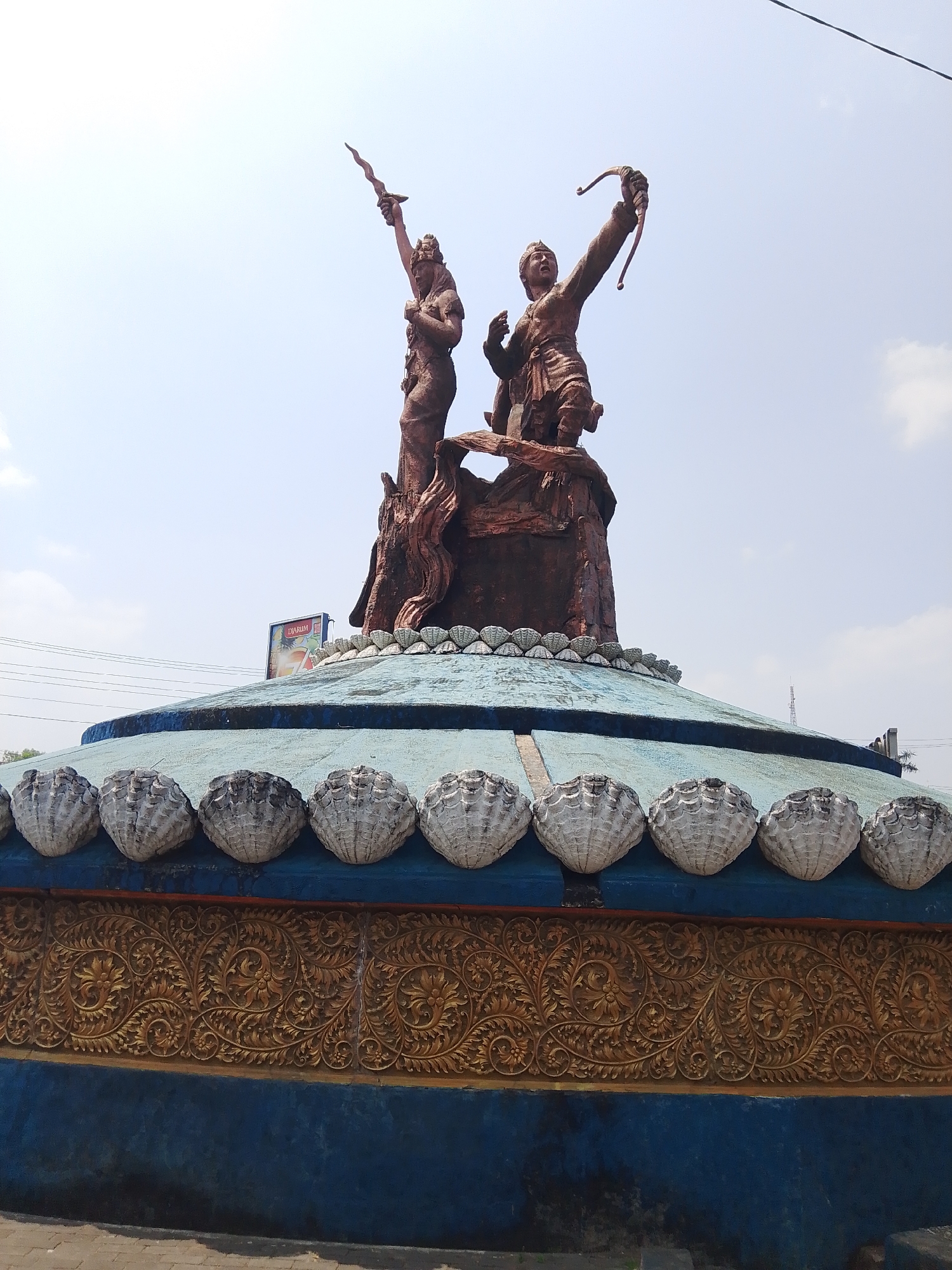 This is a medium-shot, eye-level, outdoor, daylight photograph of a statue at a roundabout in Tahunan, Indonesia. The statue is made of reddish-brown material and depicts two figures, possibly of historical or mythical significance, in dynamic poses. The figure on the left stands with its left arm raised, holding a sword or dagger aloft, while the figure on the right is depicted with a bow and arrow drawn, aiming towards the sky. The figures are mounted on a stylized, rocky base that resembles a tree trunk.
The statue is positioned on top of a circular, multi-tiered structure that forms the roundabout. The upper tier of the structure is a light blue, domed surface decorated with a row of white, shell-like ornaments along its edge. Below this is a band of intricate, golden, relief carvings featuring floral and scrollwork motifs, against a dark blue background.
In the background, a bright blue sky with scattered white clouds is visible. Power lines stretch across the upper portion of the frame. To the left of the statue, a large billboard with the "DJARUM" logo and a picture of a beverage can be seen. The foreground shows the edge of a paved area, possibly a road or sidewalk, with some shadows. The overall impression is of a public monument in a relatively open, possibly urban or semi-urban, setting.