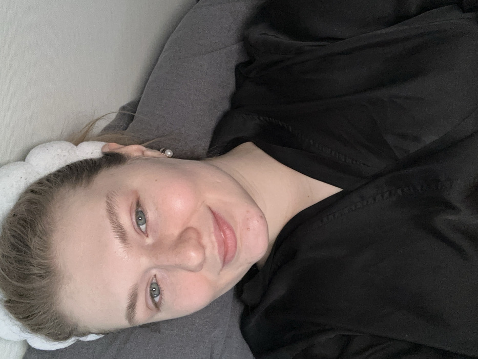 A close-up, high-angle portrait features a young adult female, appearing Caucasian, with fair skin, light blue-green eyes, and light brown or blonde hair. Her hair is pulled back from her face by a white, textured headband with distinct, uniform spherical segments. She exhibits a slight, closed-lip smile and appears to have minimal makeup, with some minor blemishes visible on her chin. She wears a black, satin or silk-like wrap-around robe and a single small, round pearl earring in her visible right earlobe. The background consists primarily of dark grey fabric, consistent with bedding or upholstery, and a lighter, plain surface in the upper right. The specific location within the city of Kazan, Russia, cannot be determined from the visual information presented.