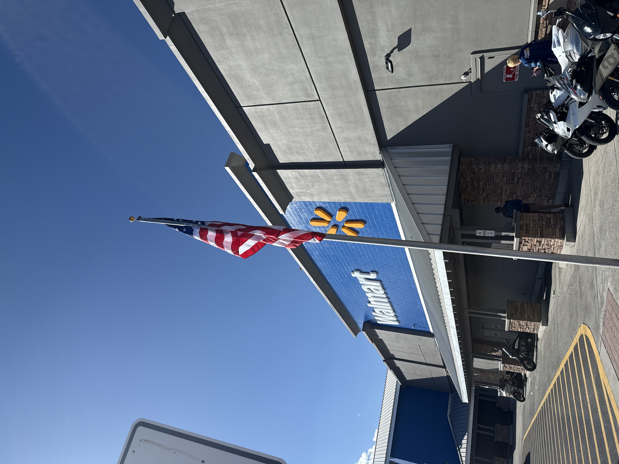 In Fernley, United States of America, an American flag waves in front of a Walmart store. The store has a distinctive blue facade with the Walmart logo prominently displayed. The exterior of the building is primarily gray with architectural detailing. Several motorcycles are parked in front of the store, suggesting a point of sale. Two individuals are visible near the entrance, one standing and the other seated. The sky is clear and blue, indicating fair weather conditions. A parking lot with yellow lane markings is in the foreground, and a portion of a white sign is visible on the left side of the frame.