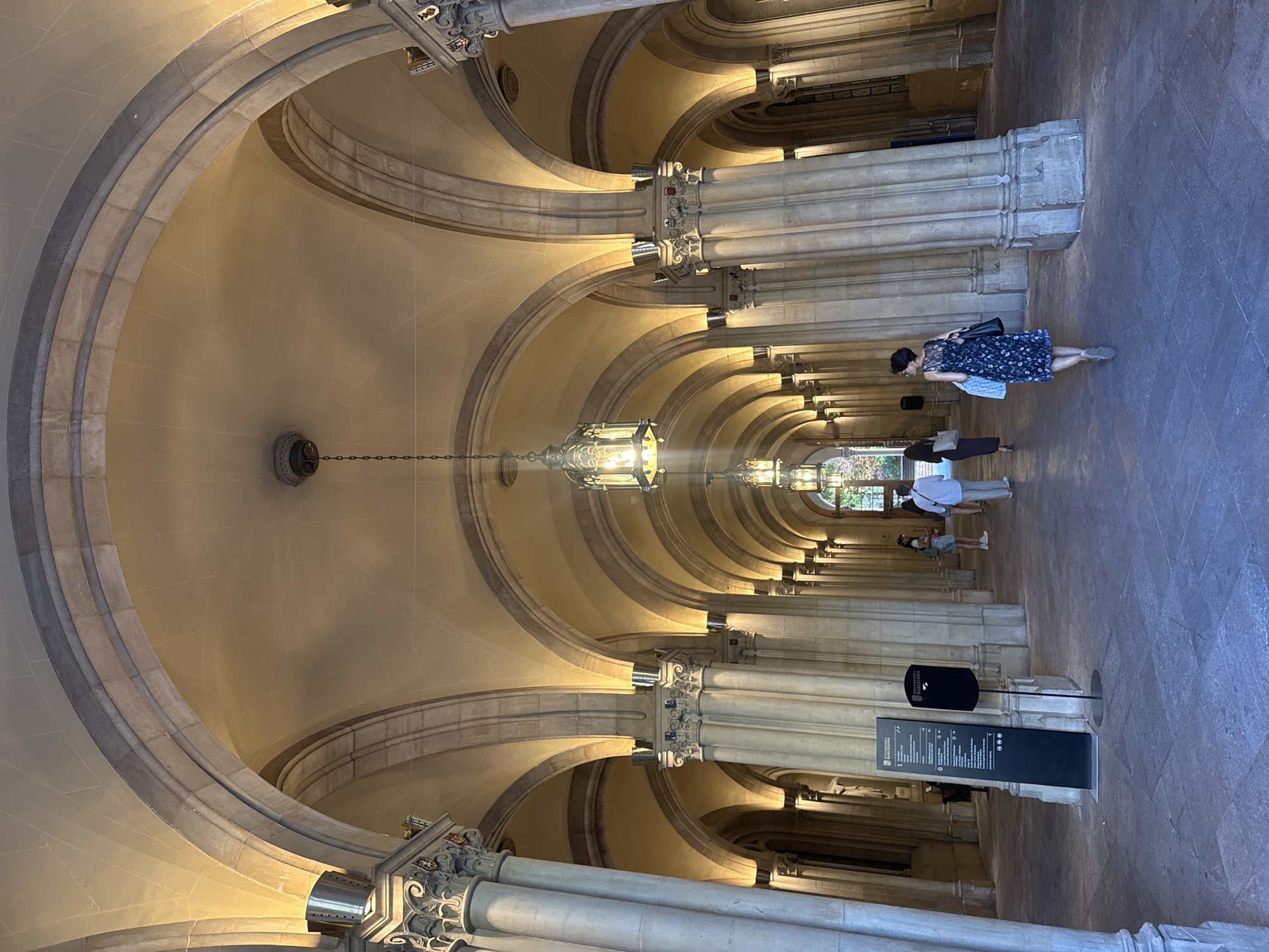 The image shows an interior view of a large hallway or corridor featuring a repeating pattern of arches and columns. The arches are constructed of stone and have a layered appearance. Hanging from the center of each arch is a decorative light fixture. Several people are walking down the corridor, including a woman in a blue floral dress. A tall, black sign or display board with writing on it is in the foreground. Based on the sign visible in the image, the location is the University of Barcelona in Barcelona, Spain.