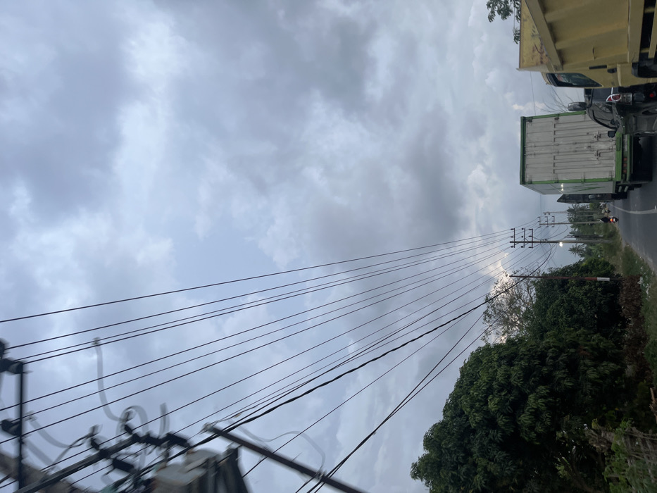 The image depicts a roadside scene in Payarangkuluh, Indonesia, under a predominantly overcast, grey sky. In the upper left, a utility pole with multiple transformers and a complex array of black electrical wires extends diagonally and horizontally across the sky, converging towards the distant horizon. The asphalt road, which appears to be a two-lane or shoulder-lined route, recedes into the background. On the left side of the road, dense green trees and foliage line the path, with some ground debris visible at their base. On the right side of the road, a line of traffic is observed moving away from the viewer. This traffic includes the rear of a yellow heavy-duty dump truck in the foreground, followed by a black SUV, and a large green and white box truck further down the road. A lone individual is operating a motorcycle on the left side of the road, also traveling away from the viewer, positioned between the trees and the moving trucks. Additional utility poles with multiple cross-arms are visible along the right side of the road in the mid-ground distance. The overall lighting is subdued due to the extensive cloud cover.