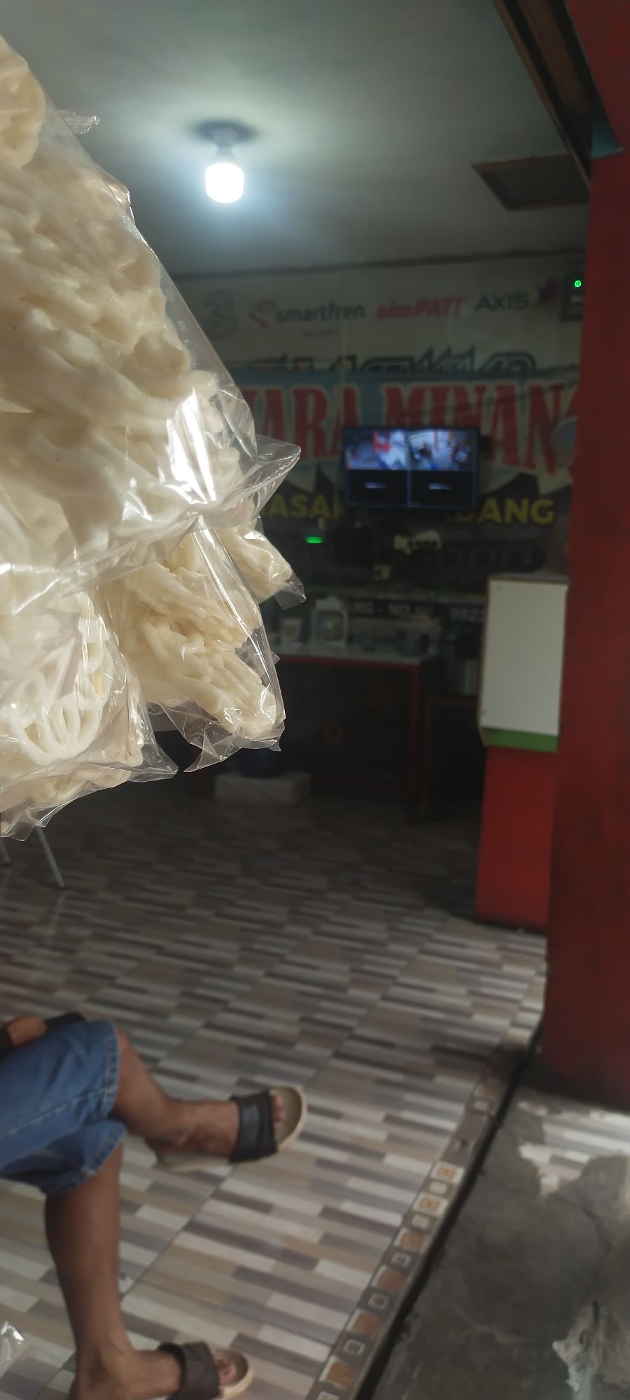 A plastic bag filled with white, circular, fried crackers is held in the foreground, obscuring part of the background. Behind the bag, a ceiling light fixture is visible. Further back, a television screen displays multiple security camera feeds. The wall behind the television features signage that includes text in Indonesian, with "SMARTFREN," "simPATI," and "AXIS" being partially legible. Below this, a larger sign reads "SARA MINANG" in red and yellow letters. The floor is tiled with a pattern of alternating light and dark brown rectangular tiles. In the lower left of the frame, the legs of a person wearing denim shorts and dark sandals are visible, with their legs crossed. The environment appears to be an indoor commercial space, possibly a shop or eatery, located in Cimaung, Indonesia.