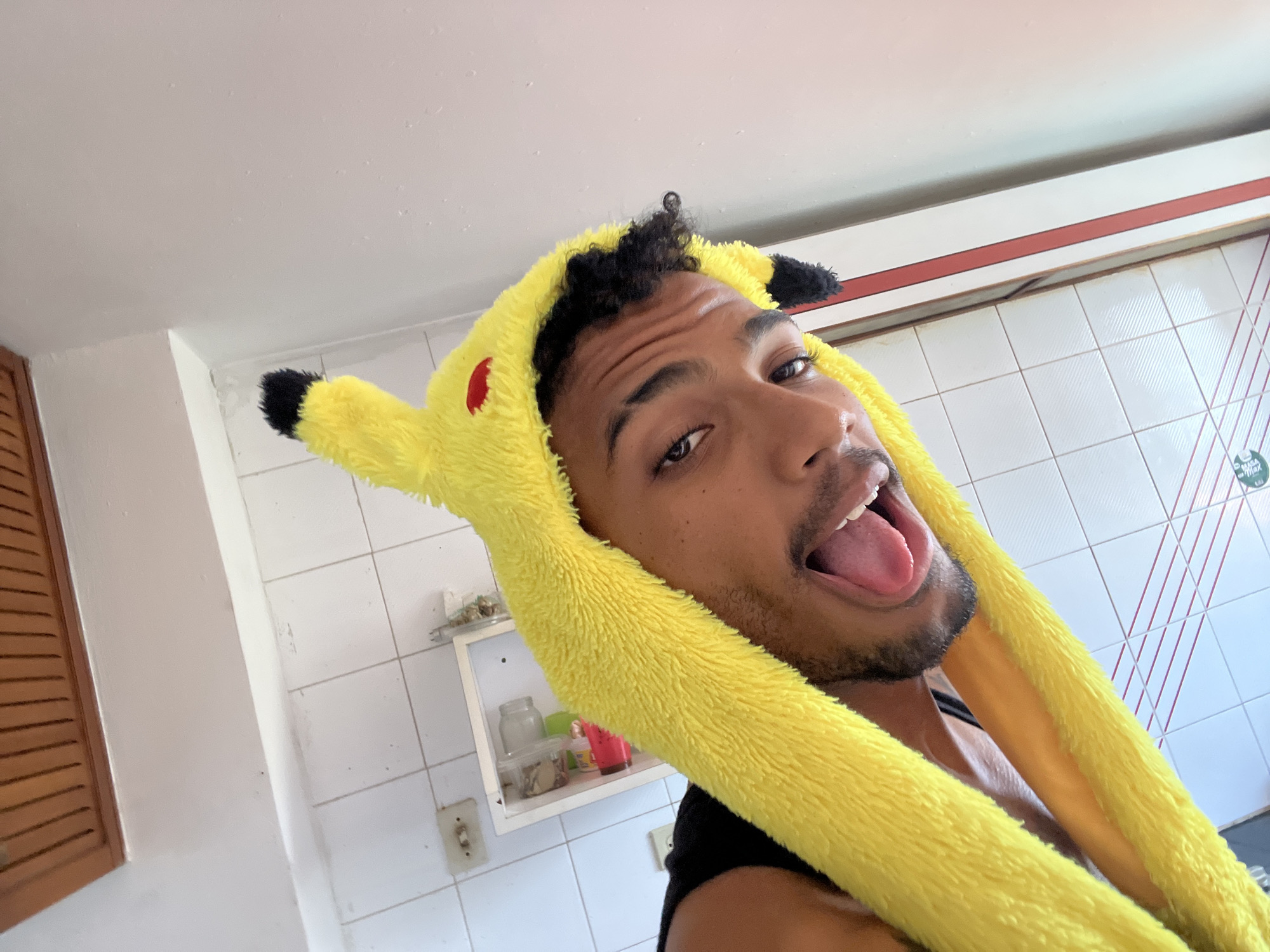 A young man with dark curly hair and a light beard is captured in a close-up selfie, holding the camera at an upward angle. He is wearing a bright yellow, plush Pikachu-themed hood, complete with black-tipped ears and red cheek accents, covering the top and sides of his head. His expression is playful, with his mouth open and tongue extended. He is wearing a black tank top.
The background reveals an indoor setting, likely a home in Porlamar, Venezuela, characterized by white tiled walls and a white ceiling. To the left, a brown wooden slatted door or cabinet is visible. A white wall-mounted shelf holding various jars and containers is present, along with light switches on the wall. A red horizontal trim accents the top of the tiled section. On a sticker affixed to the tiled wall, partial text reading "MASA" and "MAX" is discernible. The bright, natural light suggests it is daytime. The overall mood conveyed is humorous and lighthearted.