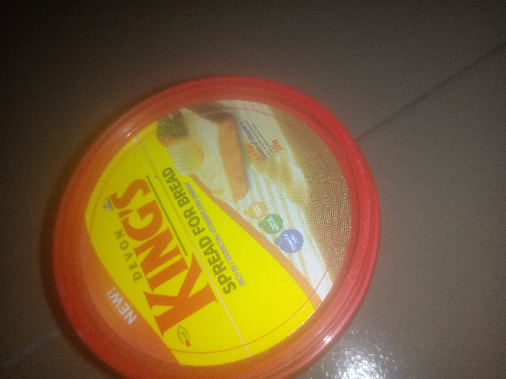 A red-lidded tub of "Kings" brand spread for bread is captured in an overhead, angled shot. The tub is primarily yellow with orange lettering and accents. The lid features an image of buttered toast. Text on the tub reads "DEVON KINGS," "SPREAD FOR BREAD," "RICH IN 7 ESSENTIAL VITAMINS AND ENERGY," and "NEW!" In smaller print, it also states "LOW FAT" and "SOURCE OF VITAMIN A." Three small circles with icons inside are visible: one green with what appears to be a leaf, one yellow with what looks like a sun, and one purple with the word "FREE." The tub rests on a textured, dark surface that appears to be linoleum or concrete. The lighting in the image is dim, casting shadows and creating some glare on the tub's surface. No people or specific city locations are depicted.