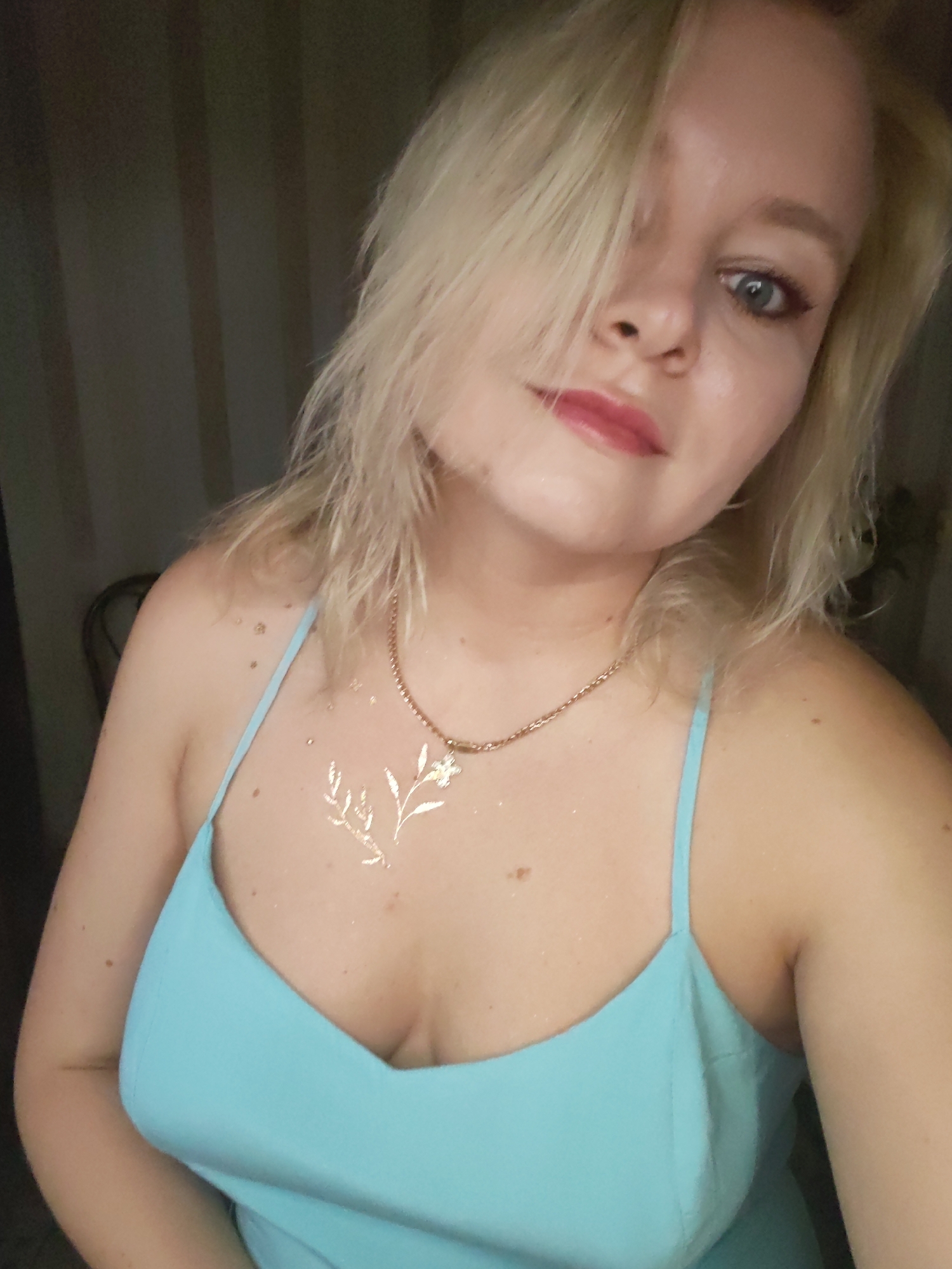 A blonde woman with light blue eyes and red lipstick is pictured in a close-up selfie. She is wearing a light blue spaghetti strap dress and a gold necklace. There is a gold floral tattoo on her chest. The background appears to be a dimly lit room with vertical blinds. The image was taken in Izhevsk, Russia.
