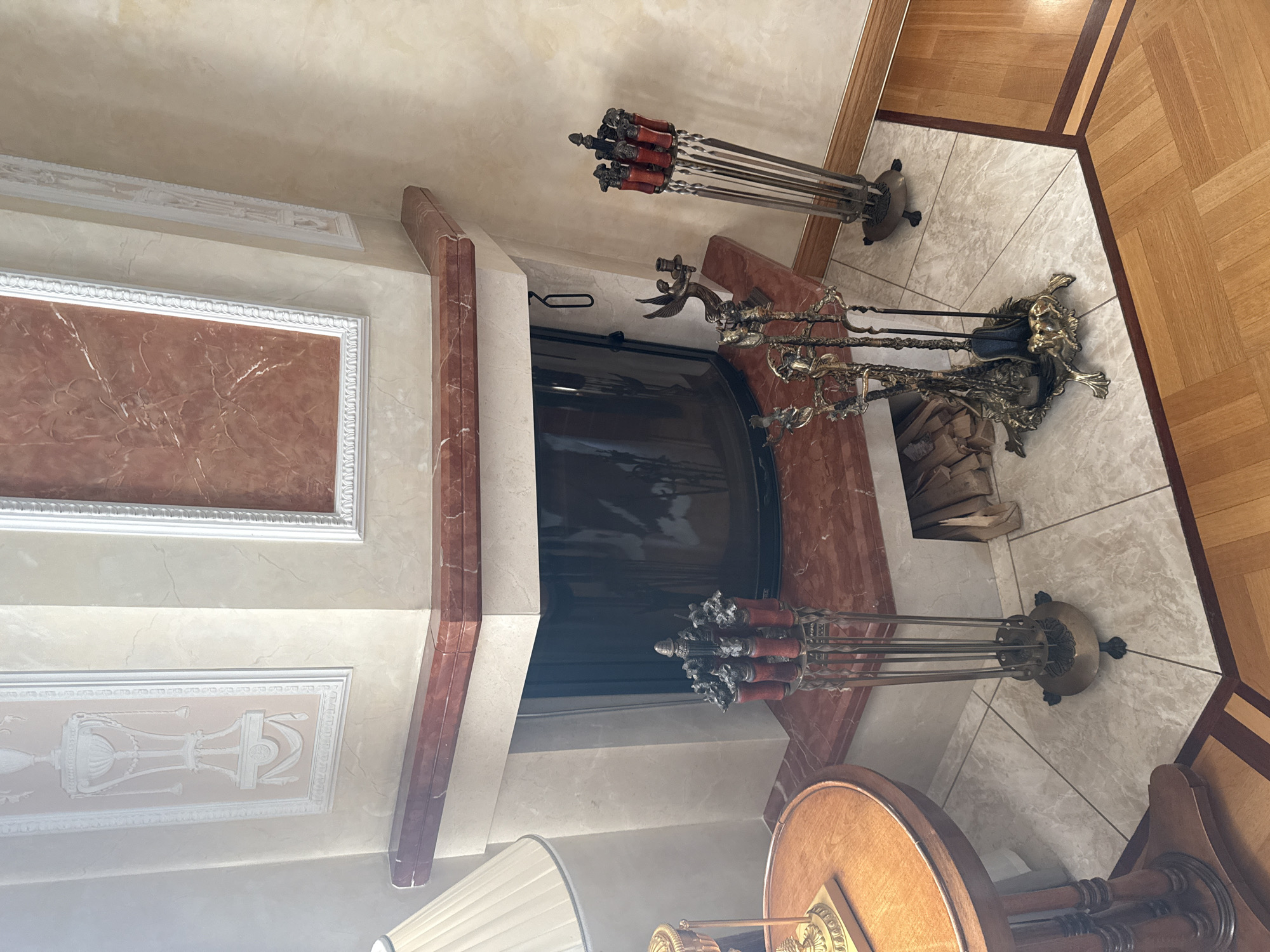The image shows an interior scene with a fireplace. The fireplace has a marble mantelpiece with a decorative red marble inlay. To the left of the fireplace, a table with a wooden top and decorative metal base is visible, with a lamp on top. In front of the fireplace, there are several fire tools, including a set of tongs, a shovel, and a poker, displayed in a decorative stand. Another set of fire tools is visible to the right, also in a stand. The floor is tiled with a marble-patterned tile. The walls are decorated with ornate plasterwork. To the right, a section of wooden flooring is visible. The scene appears to be located in a residential building in Yukki, Russia, likely a living room.