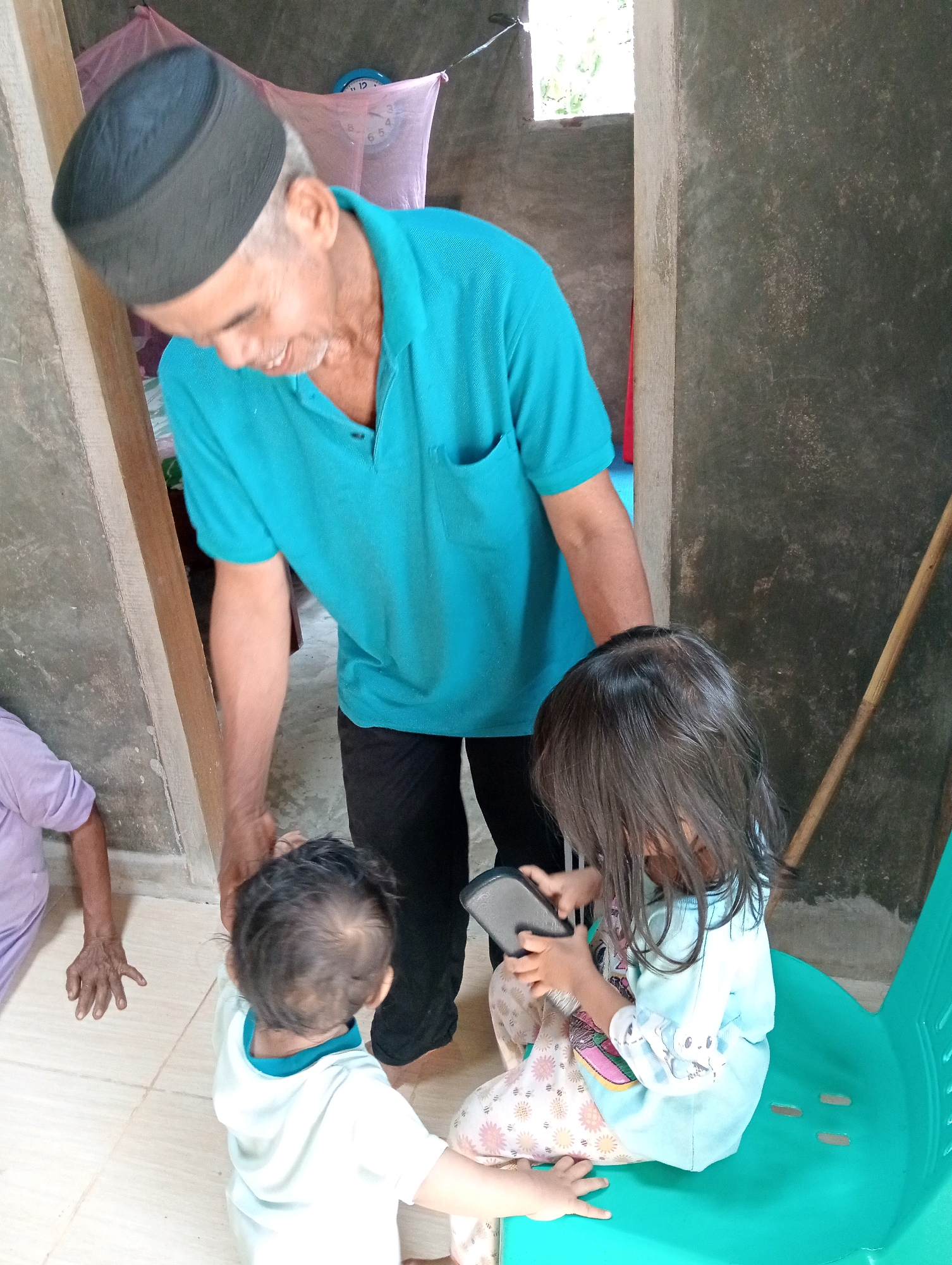 In Campago Barat, Indonesia, a man wearing a black cap and a turquoise polo shirt bends down to interact with two children. One child, a toddler wearing a white hooded shirt, stands facing away from the camera. The other child, a young girl with long dark hair, is seated on a turquoise plastic chair and holds a dark-colored rectangular electronic device, possibly a phone. She is wearing a light blue long-sleeved shirt and patterned pants. Another person, only partially visible, sits on the floor to the left wearing a light purple garment. The environment appears to be indoors, with concrete walls and a tiled floor. A wooden plank leans against a wall on the left. Behind the man, a pink fabric hangs with what appears to be a clock face visible on it.