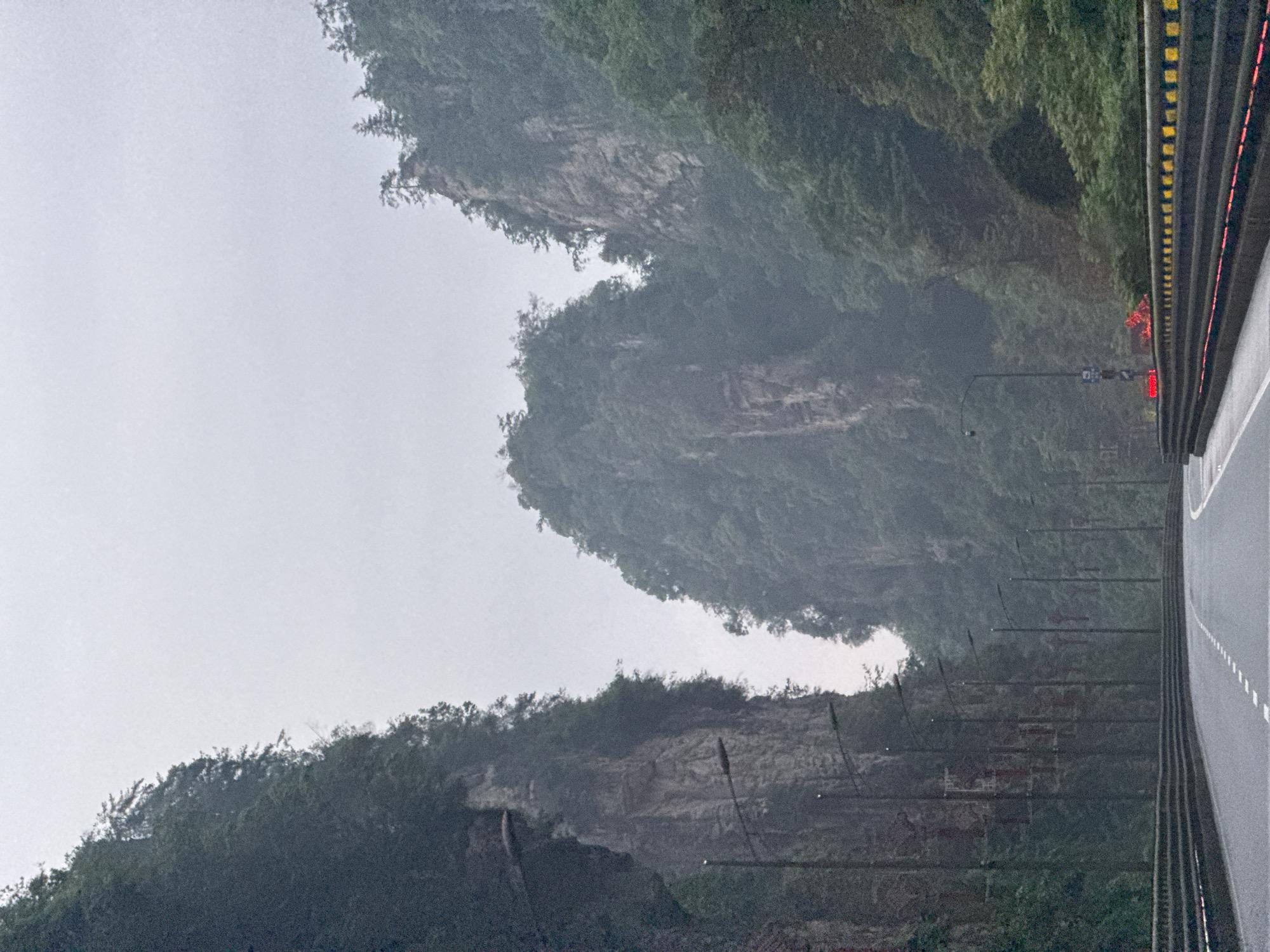 The image depicts a roadway running alongside steep, forested cliffs, likely in the Wulingyuan area of China. The road has a barrier on one side, with yellow and black markers. Above the road are cliff faces densely covered with greenery. Several light posts line the road. Traffic lights are visible in the distance. The sky is overcast. The scene appears to be a landscape shot from the perspective of a vehicle on the road.