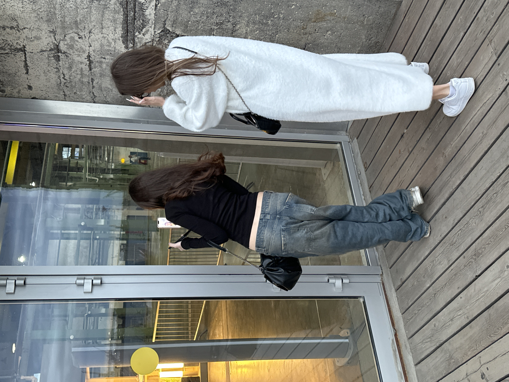 Two young women are standing on a wooden deck in front of a large glass door. The woman on the left is wearing a black long-sleeved shirt, baggy blue jeans, and white sneakers. She is holding a black purse and a smartphone, which she is using to take a photo. The woman on the right is wearing a long, white fuzzy coat, black pants, and white sneakers. She is holding a black purse and appears to be adjusting her hair. The glass door reflects the interior of a building and the exterior environment. The building has a rough, concrete wall to the right of the door. The overall scene suggests a casual outdoor gathering or outing. The specific location in Saint Petersburg, Russia cannot be determined from the visual information provided.