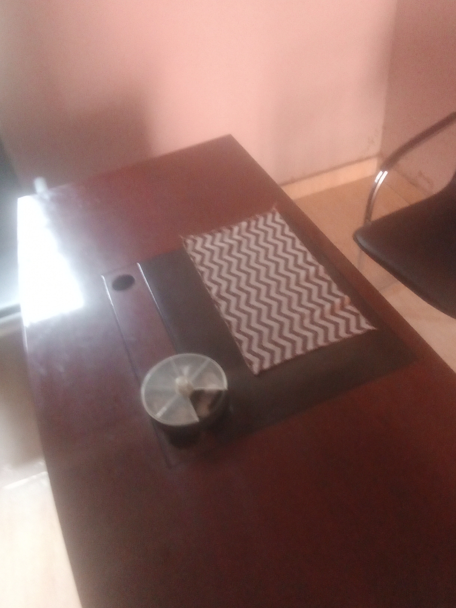 The media file shows a close-up, angled shot of a dark brown, rectangular table. On the table, there is a light-colored placemat with a zig-zag pattern in brown and white. To the left of the placemat, there's a small, round, clear plastic container with a light-colored lid, holding some brown material. The background is a softly lit, pinkish-beige wall and a glimpse of a light-colored floor. A dark chair with chrome legs is partially visible on the right side of the frame. The lighting suggests an indoor setting, possibly in the daytime due to the bright reflection on the table's surface. There is no visible text in the media file. The scene appears to be a static, still-life composition.