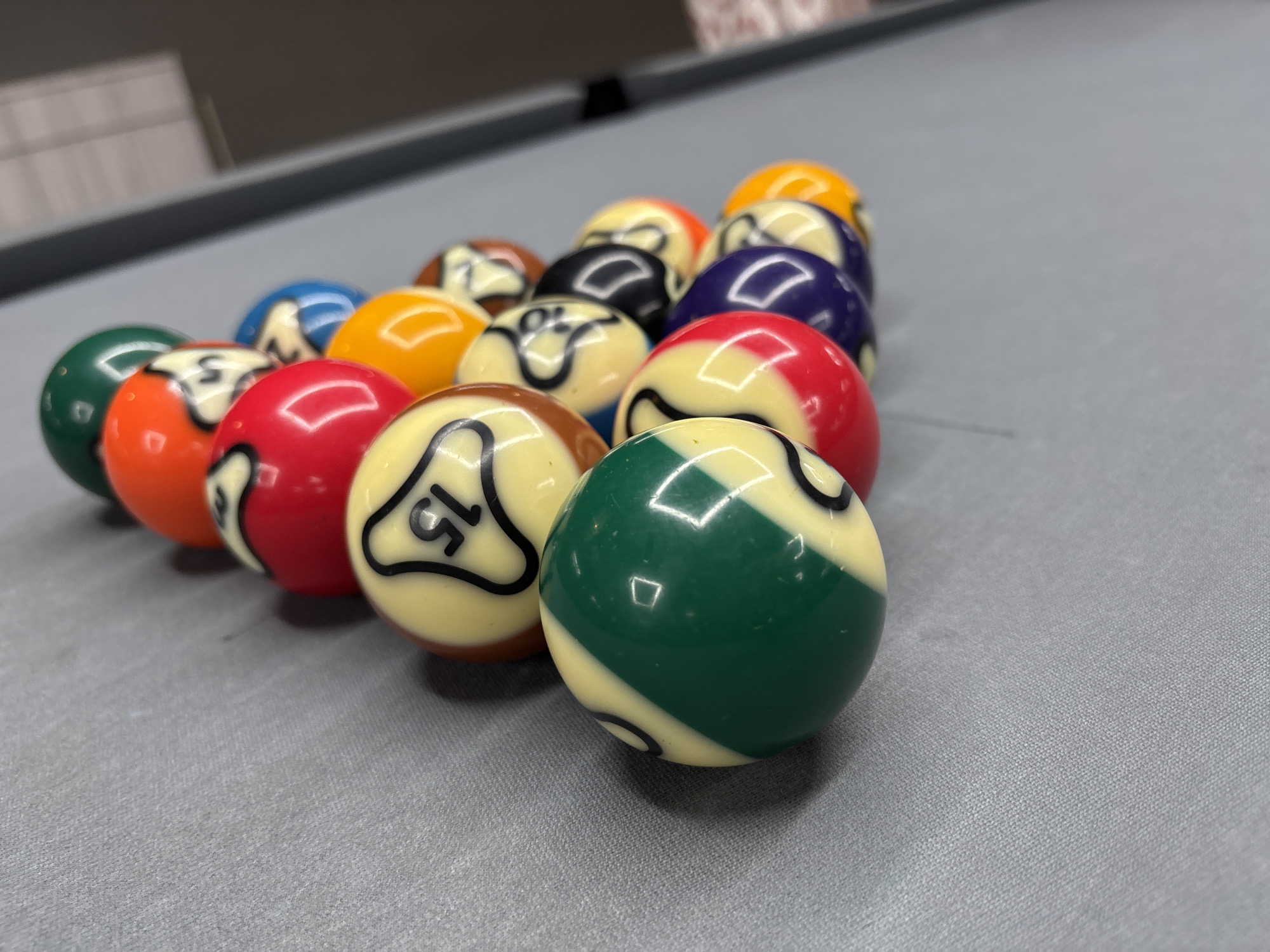 A rack of billiard balls is positioned on a gray felt table. The balls are arranged in a triangular formation, with one ball at the front. The balls include a solid green and yellow striped ball at the very front, followed by a cream-colored ball with the number 15 in black, a red and yellow striped ball, a solid red ball, a cream-colored ball with black markings, a solid orange ball, a blue ball, a solid purple ball, a black ball, a cream-colored ball with black markings, a solid yellow ball, and a cream-colored ball with black markings, and a cream-colored ball with black markings. The scene is indoors, with the background blurred and indistinct, showing what appears to be furniture or a wall. There are no people or actions depicted in the image. The location in Kuta Selatan, Indonesia is not evident from the image itself.
