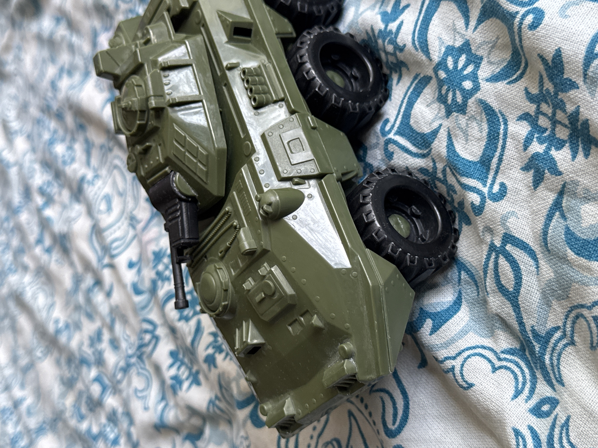 This is a medium shot, high-angle photograph of a green toy military vehicle lying on its side on a white and blue patterned cloth. The vehicle is detailed, with visible wheels, a turret with a gun, and various panels and vents. The cloth has a repeating ornate pattern of blue shapes and lines, resembling floral or paisley motifs. The lighting is soft, suggesting an indoor setting, possibly a bedroom or living area. There are no people visible in the frame. The toy appears to be plastic, with a matte finish. The overall impression is that of a simple, everyday scene, with the toy vehicle as the sole focus against a decorative background. The scene is static, with no activity taking place. The time of day and weather are not discernible. No text is visible in the image.