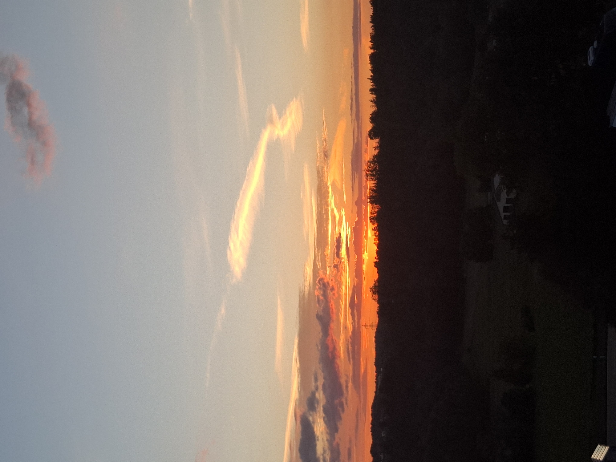 The image captures a vibrant sunset over a rural landscape near Wangen im Allgäu, Germany. The sky is ablaze with fiery oranges, reds, and yellows, transitioning to softer pinks and blues towards the zenith. Wispy clouds streak across the sky, some catching the sunset's glow. A solitary power line tower is visible on the horizon, silhouetted against the dramatic sky. Below, a dark silhouette of trees and rolling hills obscures the ground, hinting at a forested or hilly terrain. A few indistinct structures, possibly houses or barns, are visible nestled within the darkened landscape. The overall impression is one of natural beauty and tranquility at the close of day.