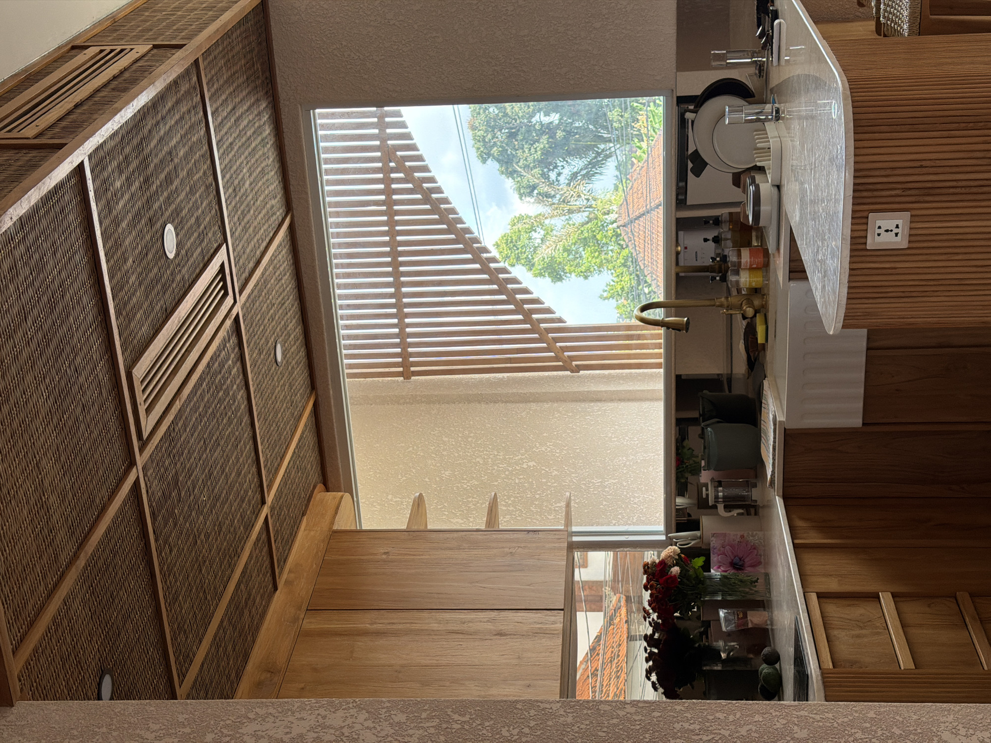 The image depicts an interior kitchen setting in Mengwi, Indonesia. The composition includes a textured wall covering with integrated light fixtures on the left. A large window with horizontal blinds reveals an exterior view of trees and a building. Kitchen elements include a countertop with a sink, dishes, and other kitchen utensils. The overall orientation is tilted, giving a skewed perspective of the scene. Additional wooden elements form the rest of the visible interior.