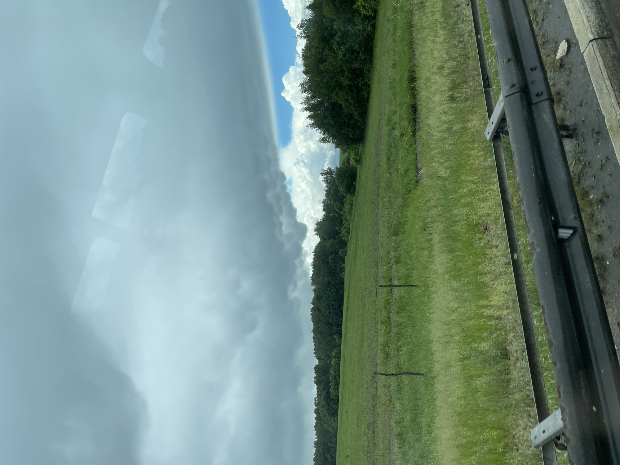 A roadside view presents a stark contrast in weather conditions. To the left, heavy, dark storm clouds dominate the sky, suggesting impending rain. To the right, there are fluffy white clouds against a blue sky. A lush green field stretches towards a tree line that separates the sky from the grassy area. A guardrail, composed of metal posts and rails, runs parallel to the road, indicating a raised or sloped embankment. The overall environment is rural or suburban, with a focus on the natural landscape juxtaposed against the manufactured safety barrier. The image does not contain any people. There is no definitive confirmation that this image is related to Кокино, Russia.
