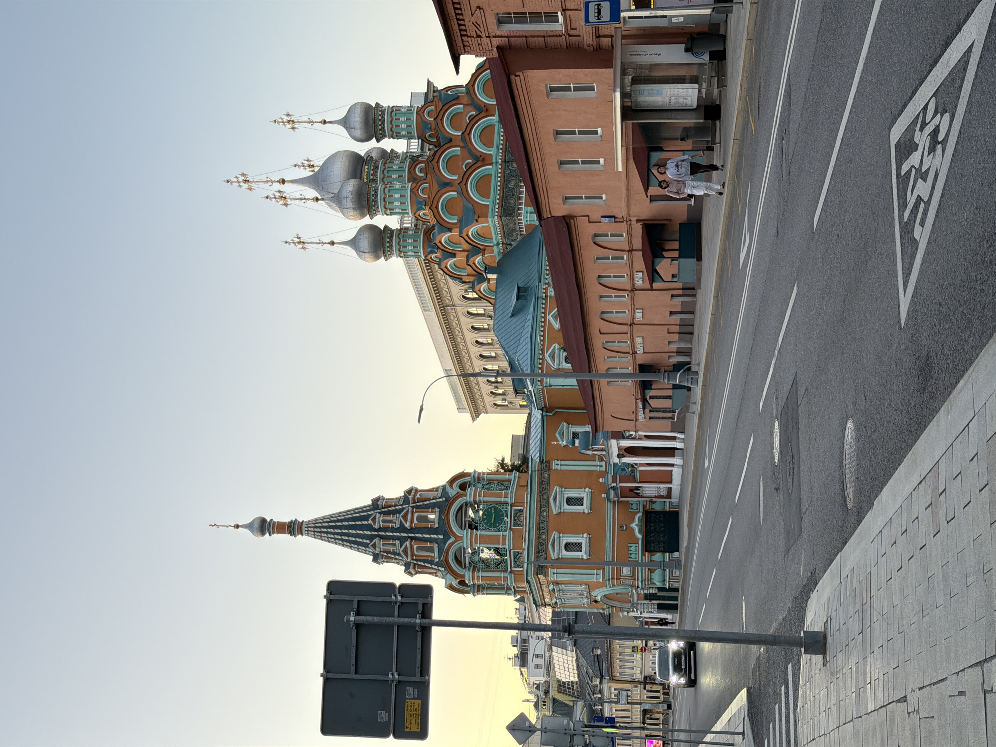 A low-angle, vertical, medium shot captures an exterior street view in Moscow, Russia, during the golden hour. The foreground features a paved sidewalk with a pedestrian crossing marked on the asphalt road. A dark-colored car is seen driving away from the camera, its headlights illuminated.
Dominating the midground are two prominent religious structures. To the left, a vibrant orange and teal bell tower stands tall, adorned with architectural details, arched windows, and a pointed spire topped with a cross. Its façade features decorative patterns and clock faces. To its right, a larger building with multiple onion domes, also in teal and orange, rises against the clear sky. The domes are capped with golden crosses.
In front of the domed structure, a long, low building with a reddish-brown brick exterior and a dark red roof stretches along the street. Several arched entrances with awnings are visible.
In the background, a modern, pale beige building with arched windows forms a backdrop to the religious architecture. Further back, other buildings and rooftops are visible, suggesting an urban environment.
Two individuals, a man and a woman, are walking along the sidewalk in front of the reddish-brown building, heading towards the right of the frame. A bus stop shelter is also present. The overall scene depicts a blend of historical and modern architecture on a city street bathed in the warm light of dusk.