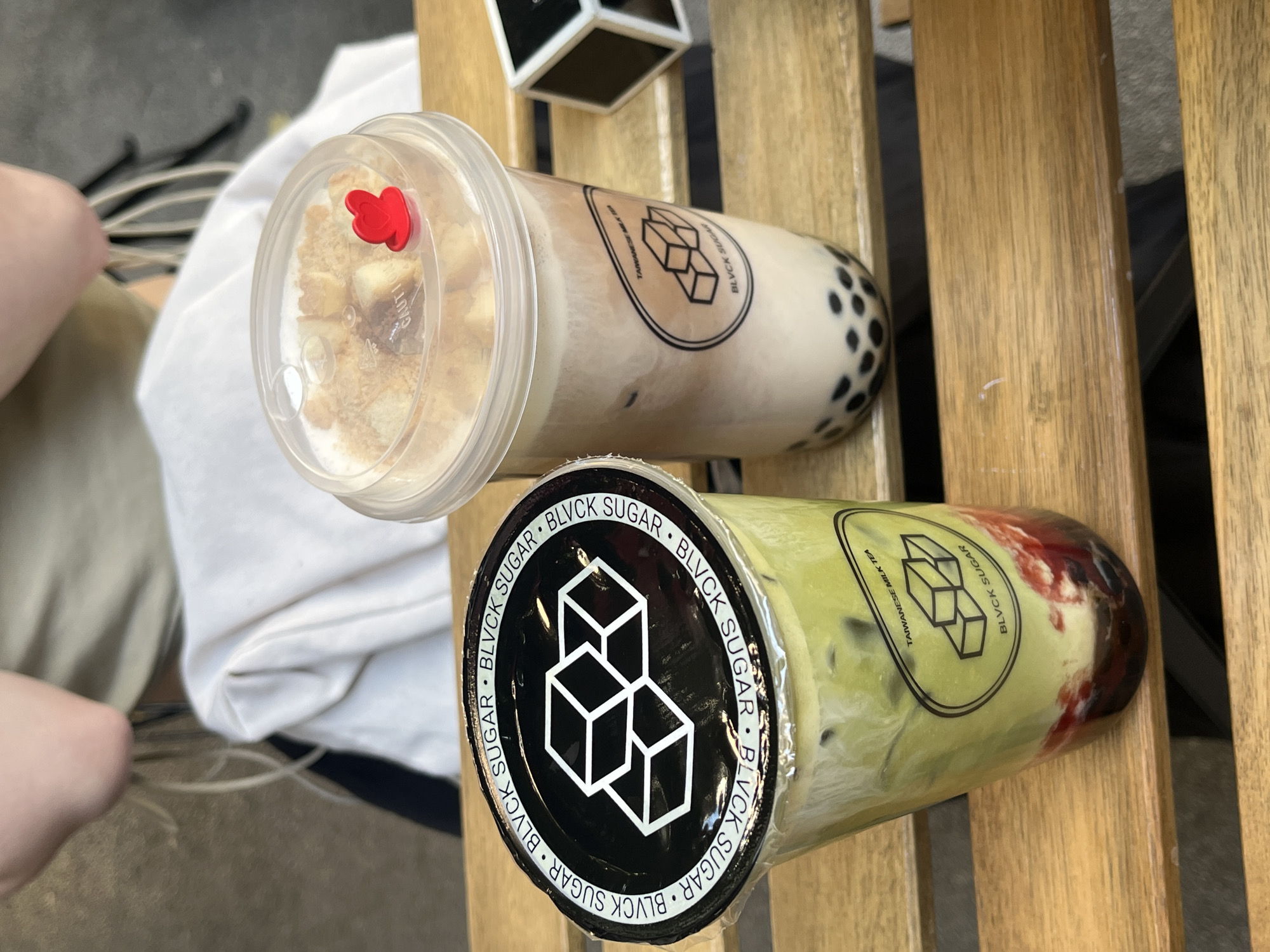 The image shows two "Blvck Sugar" Taiwanese milk tea drinks on a wooden surface. One drink is light brown with boba at the bottom and appears to have a topping of small cake pieces and a red heart-shaped object on top, while the other is green with dark red elements at the bottom. Both cups have the brand's logo prominently displayed. A person's leg is partially visible in the lower-left corner, and a small black and white box is in the background. The scene suggests an outdoor setting, possibly at a cafe or similar establishment. I cannot determine the location is in Belgrade, Serbia, from the image.