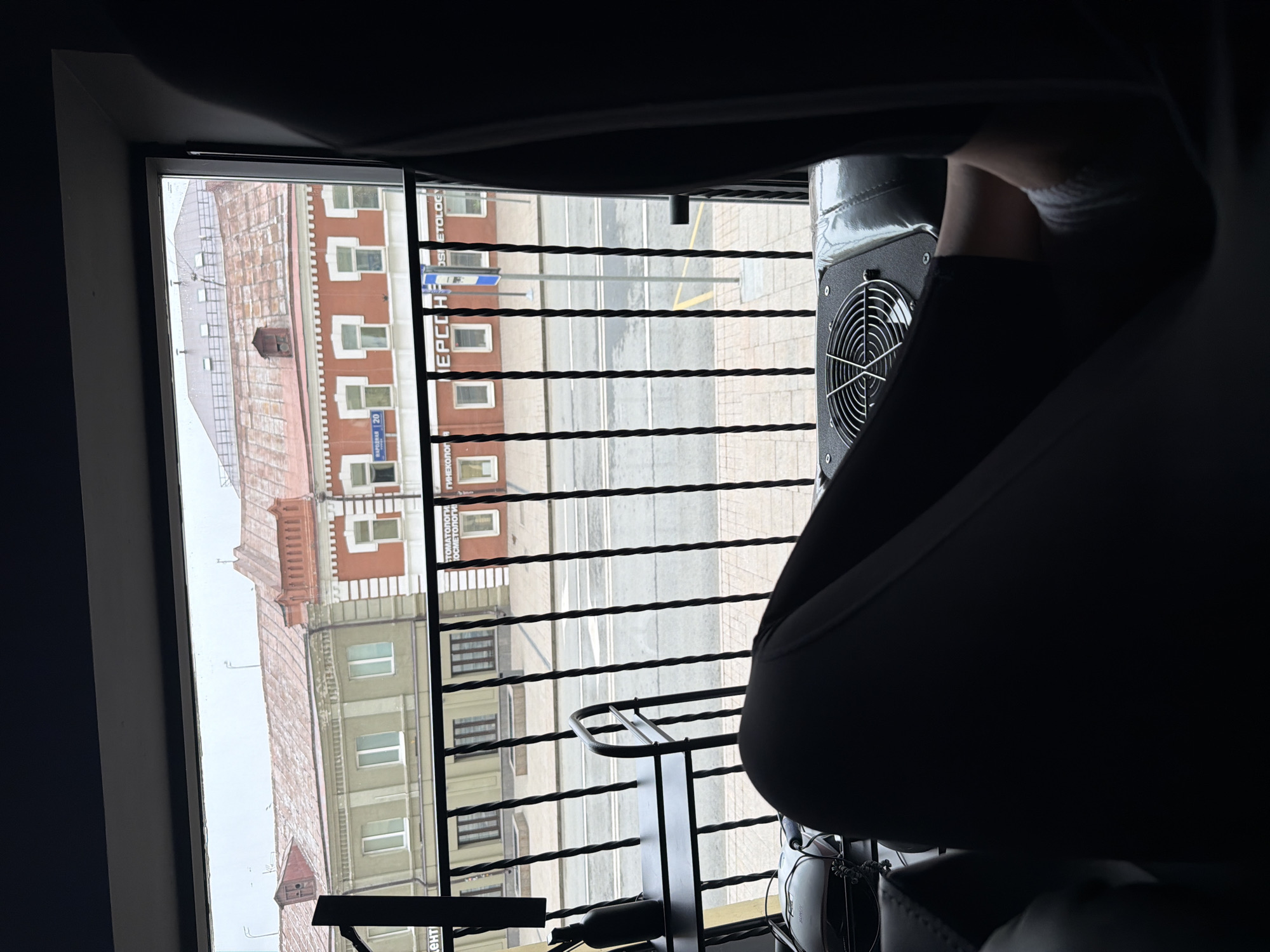 The image shows a view from a high vantage point, likely a window, overlooking a street in Moscow, Russia. Outside the window, several buildings are visible, with distinct architectural styles and colors, including brown, red, and green. A sign with Cyrillic characters, including "ЕРССН," is visible on one of the buildings. The street below is seen with a yellow centerline. A balcony rail with twisted metal posts partially obstructs the view. The foreground is dark, featuring an object with a fan, potentially a chair or equipment, and a person seated.