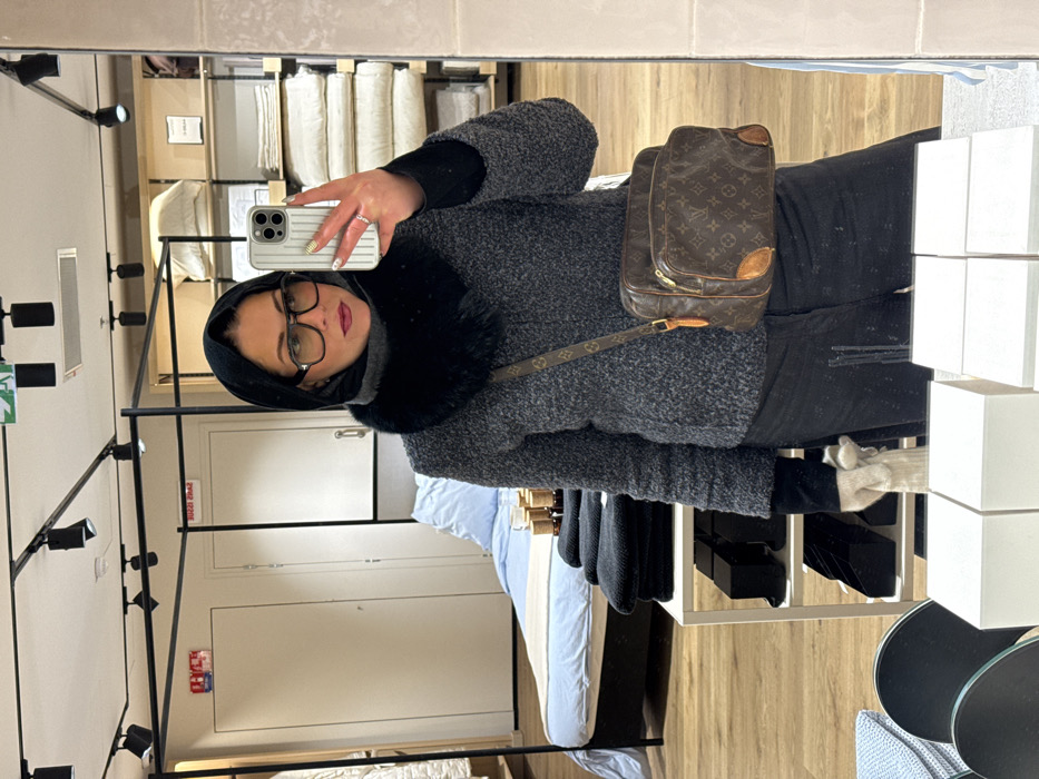 The image displays a female subject capturing a mirror selfie inside a Zara Home retail store in Paris, France. The woman is presented from the waist up, adorned in a black headscarf, a black faux-fur or fluffy neck warmer, and a textured dark grey jacket. She wears black-rimmed eyeglasses with tinted lenses and dark red lipstick. Her left hand, visible in the reflection, holds a silver-toned iPhone encased in a ribbed white or light grey cover, with a ring present on her left ring finger. A brown Louis Vuitton monogram canvas shoulder bag with leather accents is slung across her body. Her right hand, also visible in the reflection, holds a light-colored, possibly white or beige, knitted glove. The reflected background reveals various home furnishings displays typical of a retail environment. To the viewer's left, a bed with light blue sheets and a white blanket is arranged, adjacent to a white display stand holding folded dark towels and small bottles with wooden caps. To the viewer's right, shelving units are stocked with folded white and cream-colored linens. The ceiling features black track lighting fixtures and a green emergency exit sign. The floor is light-toned wood. The store name "ZARA HOME" is discernibly visible in reversed text on a white wall within the reflection.