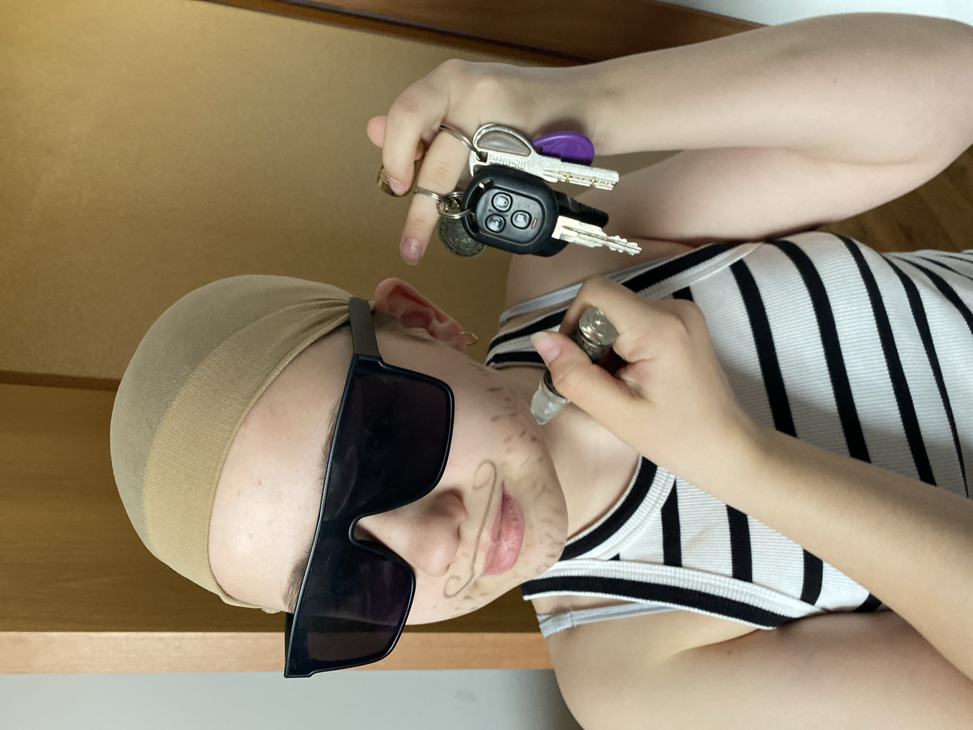 A person is depicted in an indoor setting, rotated 90 degrees clockwise. They are wearing a light brown hairnet or bald cap, large black sunglasses, and a white and black striped tank top. Their face has drawn-on facial hair, specifically a mustache and a goatee, suggesting a playful or costume-like intent.
In their left hand, the person holds a keychain with several keys, including a black car remote key with three buttons, and other metallic keys, one of which has a purple plastic cover. The word "SAMSUNG" is visible on one of the metallic keys. Their right hand holds a metallic-tipped pen or marker, positioned near their face, implying they are either in the process of drawing the facial hair or have just finished. The background consists of a light brown wall or surface and a lighter area below. The lighting suggests an indoor environment.
