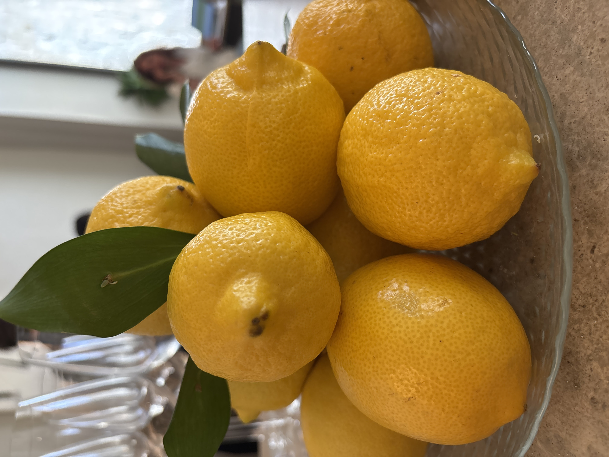 A close-up, eye-level shot shows a pile of bright yellow lemons in a clear glass bowl, situated on a textured countertop. The lemons are predominantly in focus, showcasing their bumpy rind and vibrant color. Two green leaves are strategically placed amongst the lemons, adding a touch of freshness. In the background, out of focus, are several clear wine glasses and a blurred figure of a person. The setting appears to be indoors, with natural light coming from a window on the right side, where another blurred person can be seen. The overall impression is one of a still life arrangement, possibly in a kitchen or dining area in Saint Petersburg, Russia.