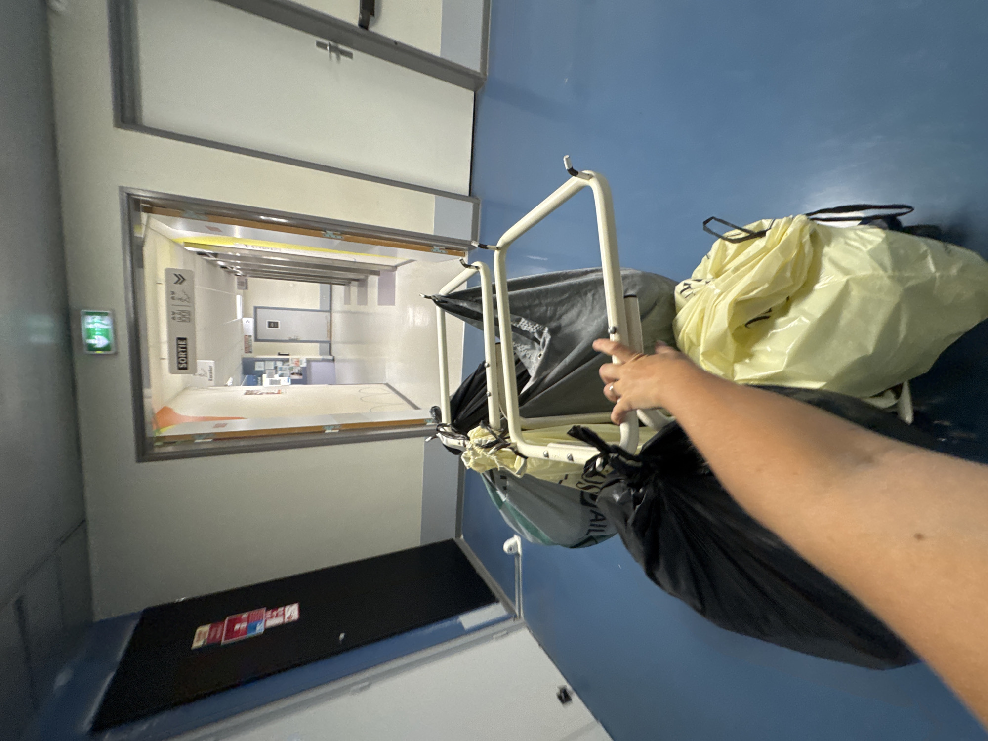 The image shows a first-person perspective of a hallway in what appears to be a healthcare facility. The hallway has blue flooring and pale walls. Several doors are visible along the right side of the hallway. In the foreground, a person's arm and hand are visible, holding onto a cart that carries several large bags. The bags are of different colors, including black, gray, and yellow. Some bags appear to be laundry bags, while others may contain waste. An illuminated green "EXIT" sign is visible on the ceiling in the distance, along with a "SORTIE" sign indicating an exit. The overall scene suggests a functional environment within a building, likely a hospital or clinic, where waste or laundry is being transported. Tourcoing, France is a city in the Nord department of northern France, known for its industrial past and growing service sector, which includes healthcare facilities.