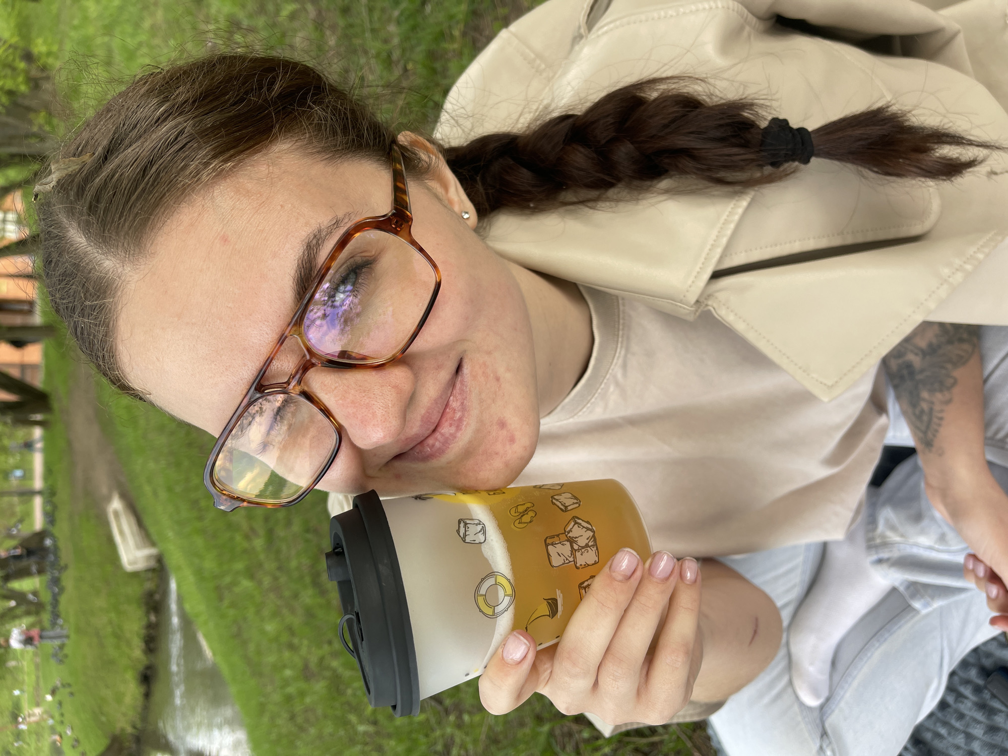 A young woman sits outdoors near a grassy area, possibly in a park. She wears glasses, a beige jacket, and light-colored jeans. Her brown hair is styled in a braid secured with a black elastic band. She holds a tumbler with a yellow beverage. A stream or small body of water is in the background. The presence of people and trees hints at a park setting, potentially in Moscow, Russia.