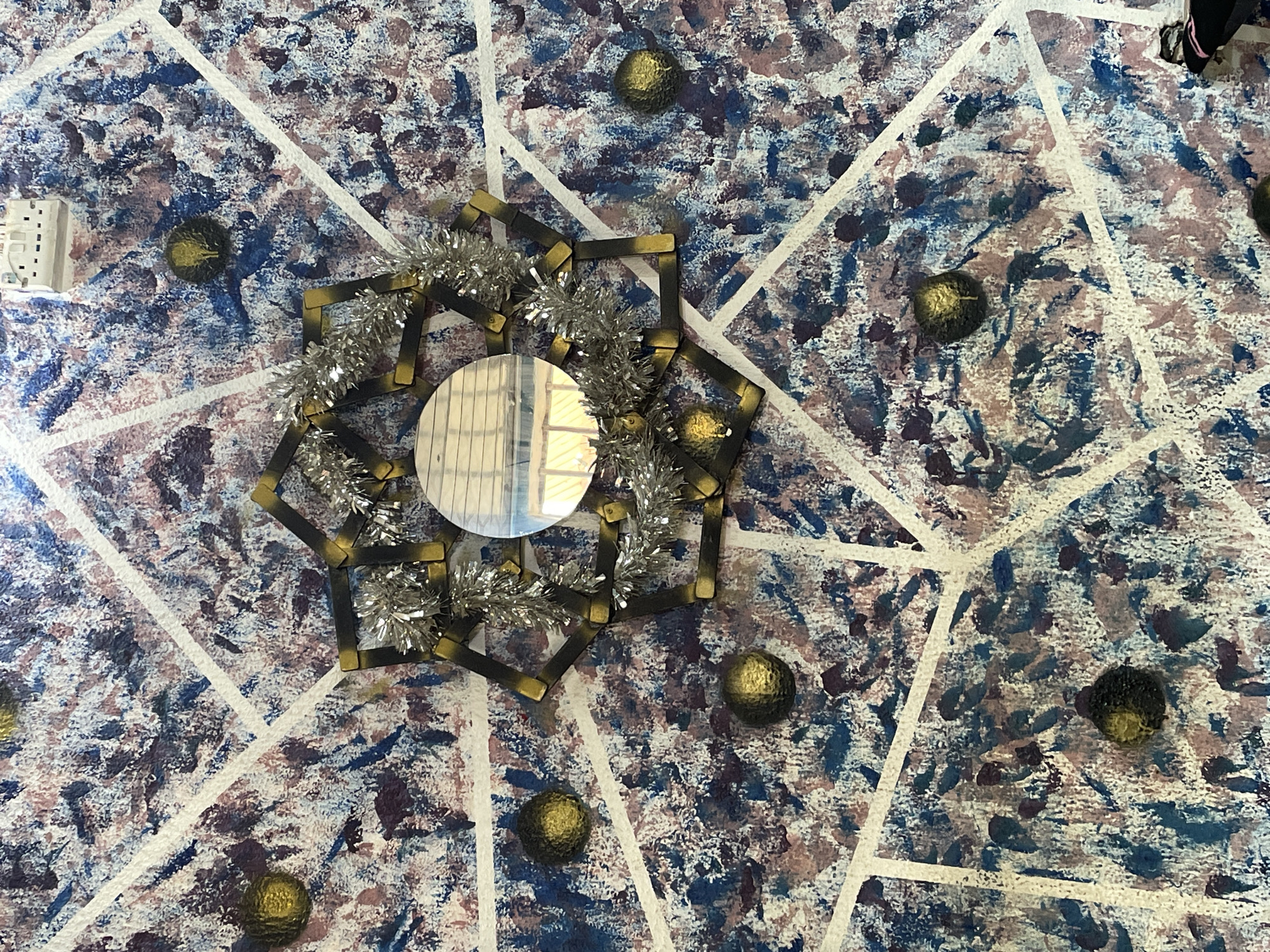 A high-angle shot shows a ceiling painted with a splattered pattern of blue, brown, and white, divided by broad white lines in a geometric, star-like pattern. At the center of the image is a circular mirror framed by a starburst of gold and black rectangular segments, adorned with silver tinsel. Scattered across the ceiling are several small, round, gold-colored objects. A white electrical outlet is visible at the upper left. The ceiling's location is in Sabon Tasha, Nigeria, but no other information is available about the room or building. No people are identifiable.