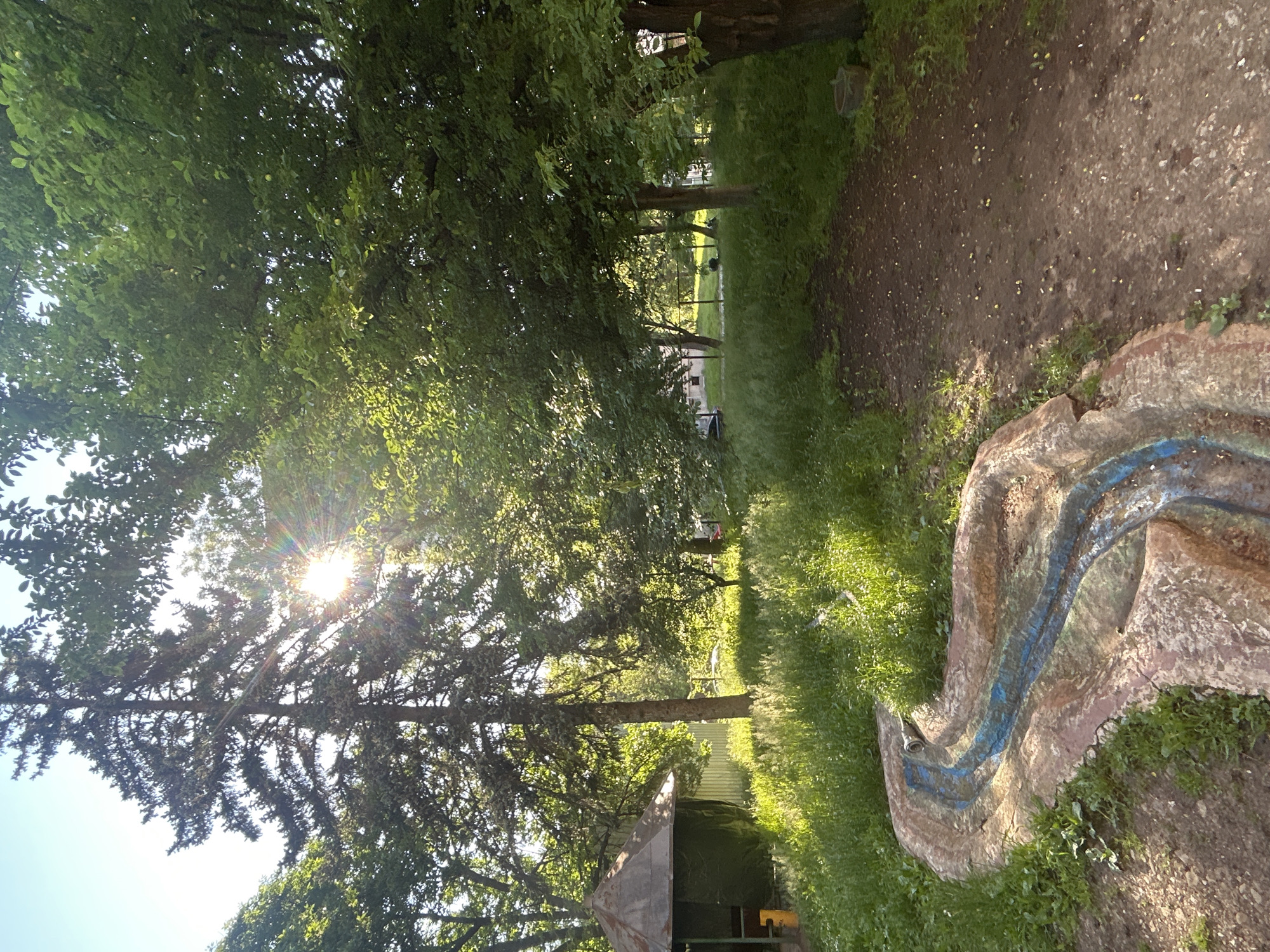 The image shows a sunlit path through a wooded area in Unknown, Unknown. Trees line the path, with sunlight filtering through their leaves. A small, unidentifiable structure is seen near the bottom of the frame. To the right, an artificial, weathered object with a blue marking sits on the grass. The ground is a mixture of dirt and grass. The path appears to lead to a grassy area in the distance.