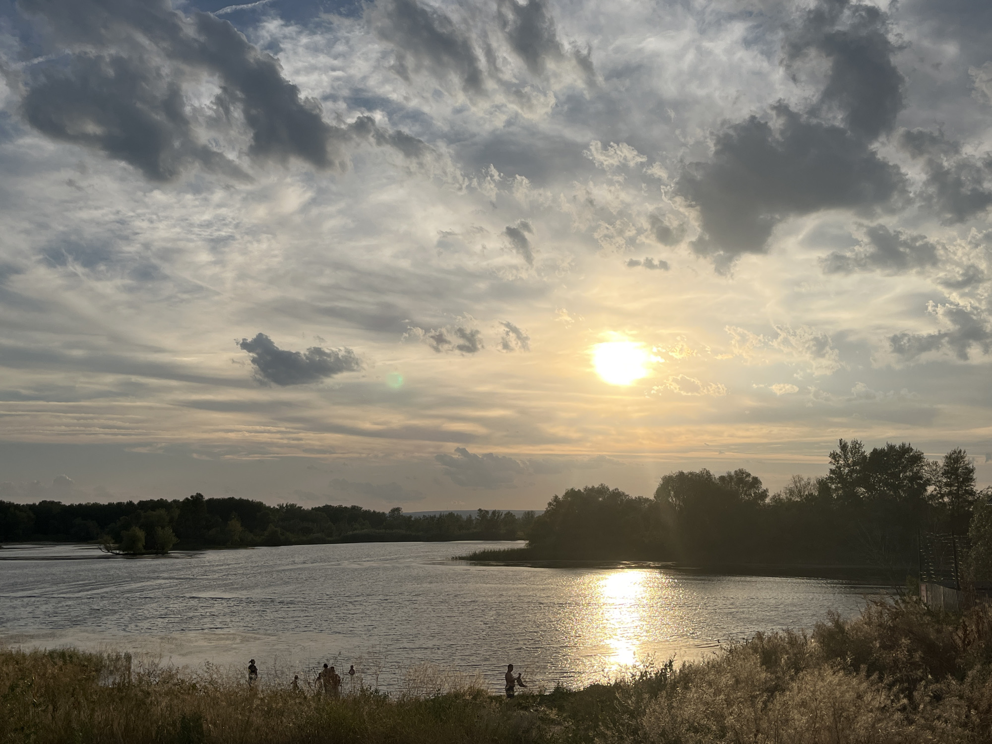 This is an image of a river during sunset, likely taken in the vicinity of the village of Шумейка, Russia. The sun is setting on the right side of the frame, casting a warm glow on the water and the sky. The sky is filled with a mix of dark and light clouds, creating a dramatic backdrop. On the far bank of the river, a dense line of trees is silhouetted against the fading light. In the foreground, tall grasses and reeds grow along the riverbank. Several figures, presumably people, are visible standing near the water's edge on the left side of the image, some appearing to be in the water. The overall scene depicts a serene natural landscape at dusk.