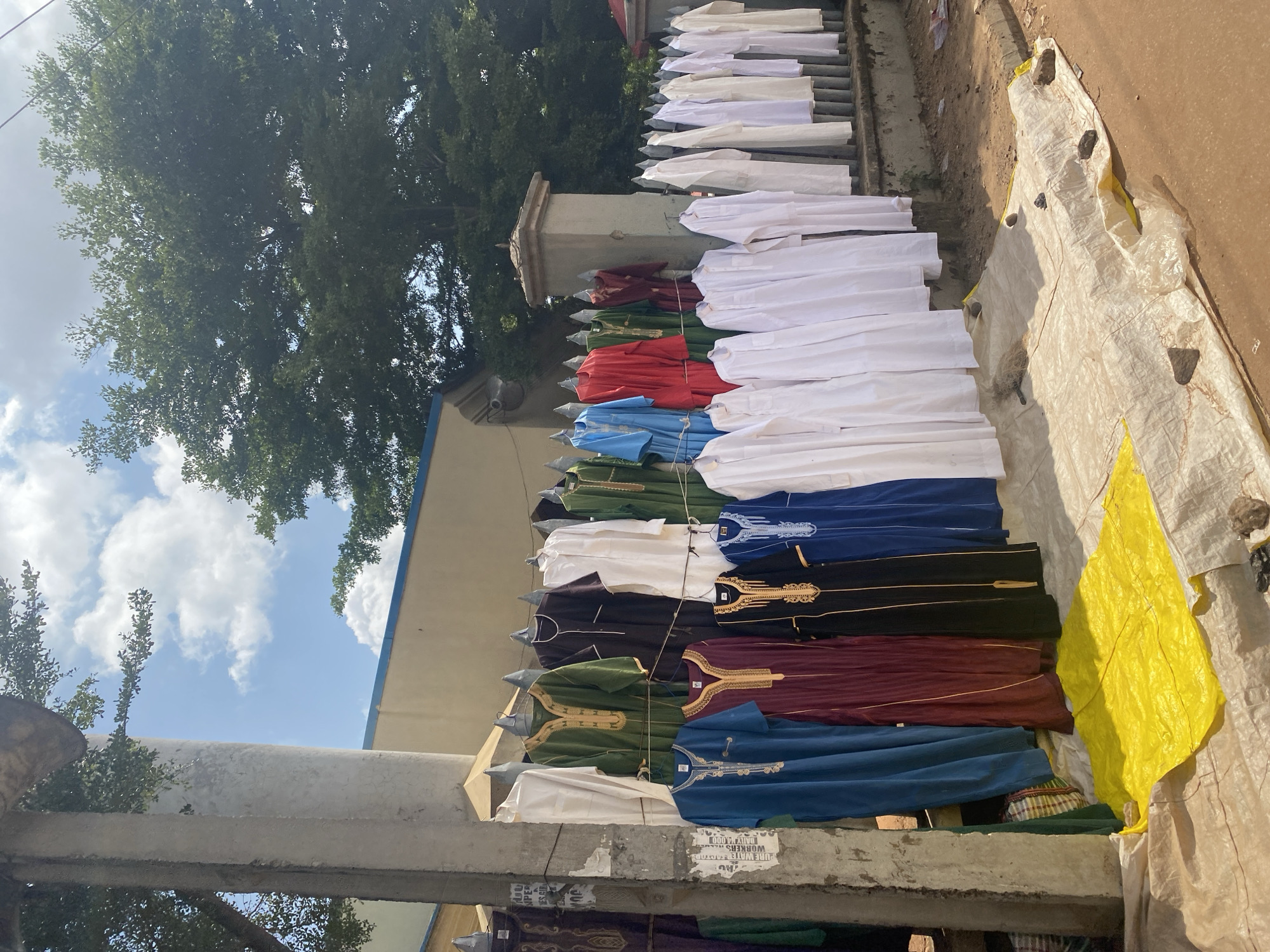 This photograph captures an outdoor clothing stall in Ado, Nigeria. The scene is dominated by a vibrant display of traditional garments, likely for sale, hanging from a fence and a pole. The clothing consists of various robes and long-sleeved shirts in an array of colors including white, blue, green, maroon, and black, some adorned with intricate gold embroidery. A bright yellow tarp is spread on the ground in the foreground, with some rocks and a broom on it. In the background, a building with a blue trim and lush green trees are visible against a clear blue sky with scattered clouds. The overall impression is of a bustling marketplace setting, typical of a Nigerian city.