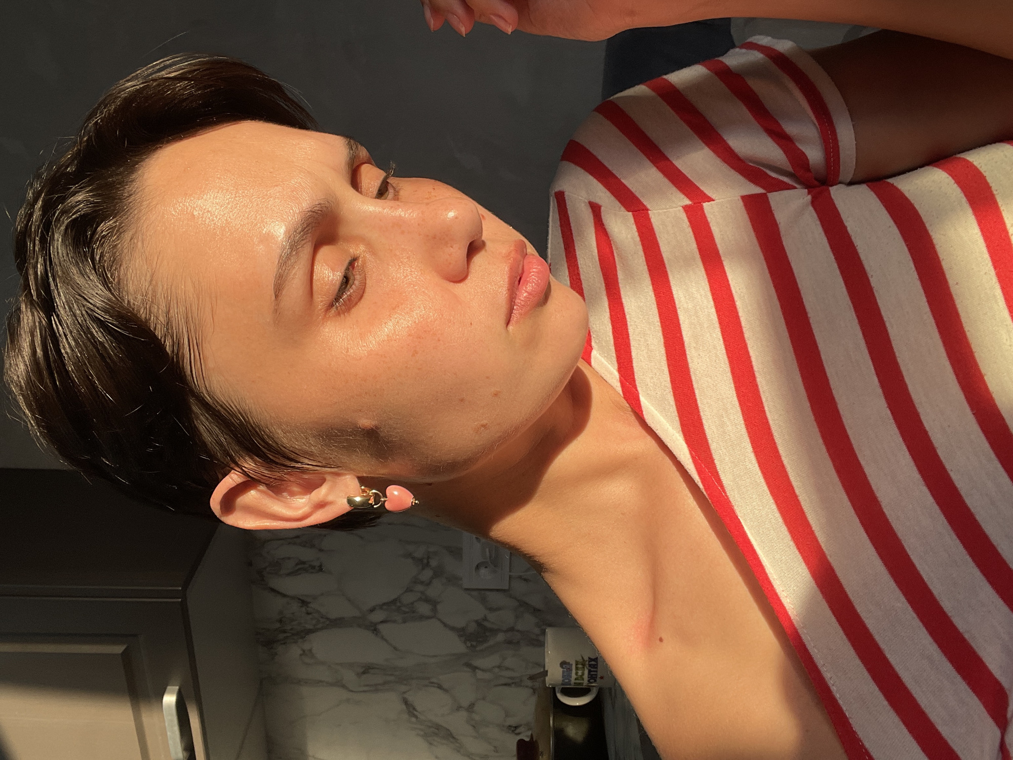 A person with short, dark hair and fair skin is illuminated by sunlight. The person's eyes are closed, and their face is angled upward. They are wearing a white and red striped shirt. A pink heart-shaped earring is visible on their left ear. The background includes a gray cabinet and a light-colored countertop. A mug with illegible text is visible on the countertop. The location is inside a building, possibly a home, but there is no indication that the location is in Izhevsk, Russia.