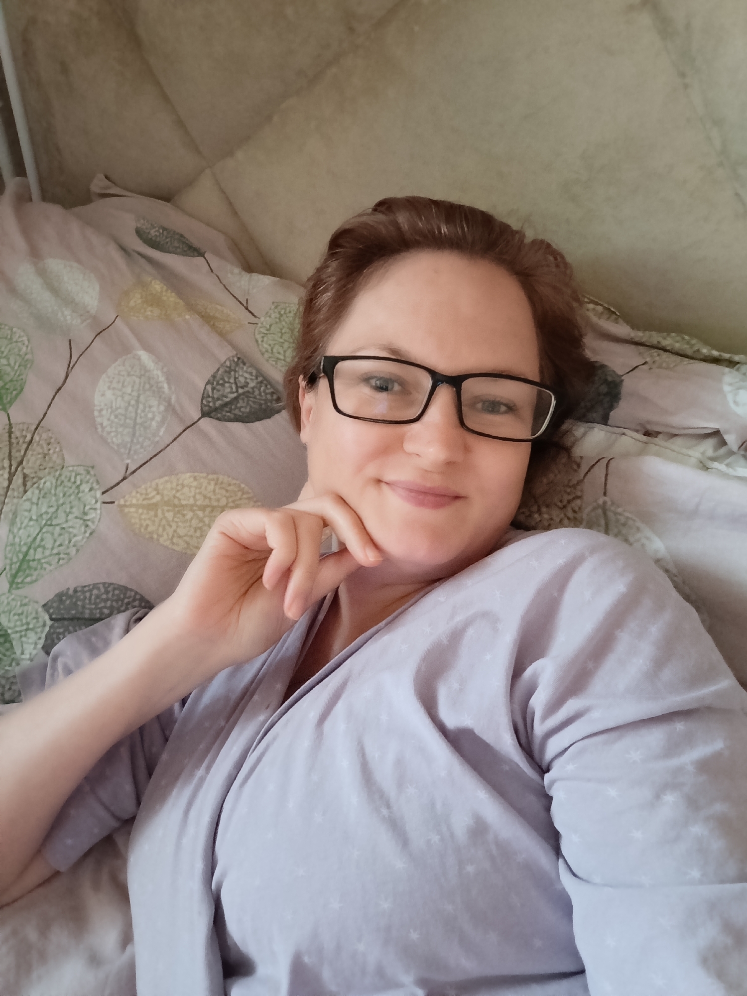 In the city of Марковский, Russia, a woman with reddish-brown hair and glasses lies in bed, propped up on pillows with a patterned duvet. She is wearing a light purple robe with a star pattern and has her right hand resting on her chin. The background consists of a lightly textured wall and the bedding. The image is a selfie taken from a slightly elevated angle.