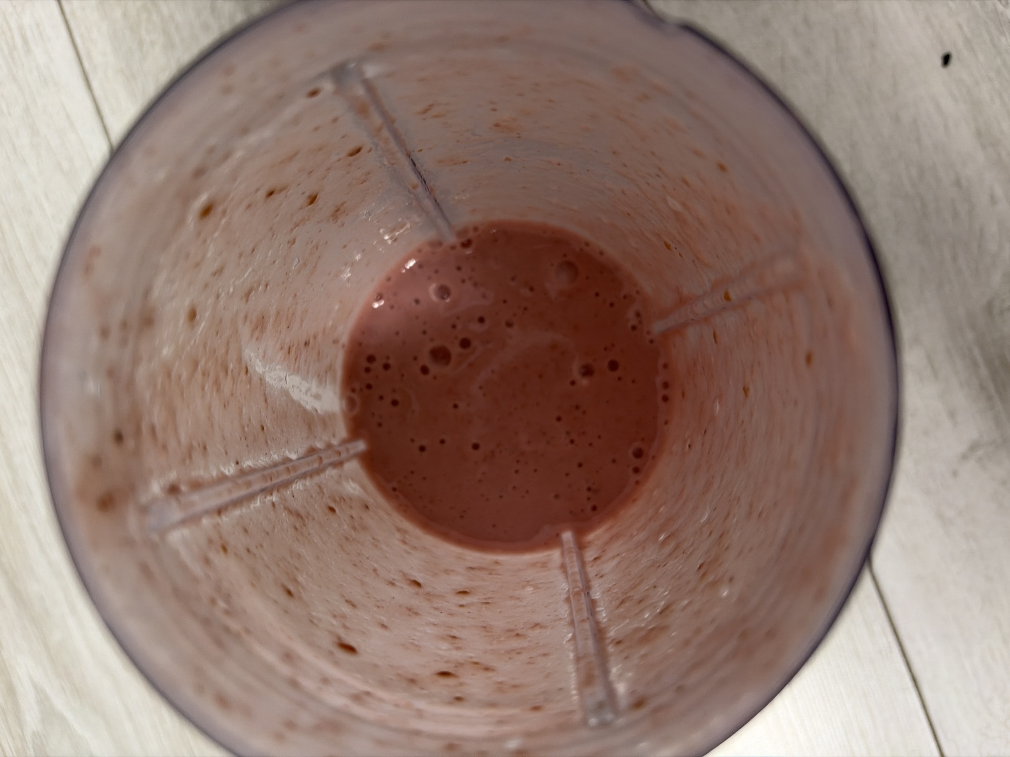 A close-up, overhead view shows the inside of a blender pitcher with pink smoothie residue clinging to the sides. The smoothie itself is a lighter pink, with small bubbles visible on the surface. The blender pitcher is translucent purple and sits on a light-colored wooden surface, possibly a table or countertop. The scene appears to be indoors, in a domestic setting. There are no people or active processes depicted.