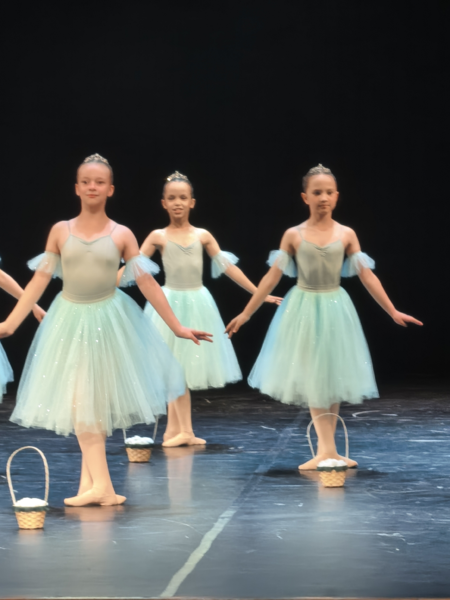 The image shows three young female ballet dancers on a stage in front of a black backdrop. The dancers are wearing matching light blue tutus and leotards, and each has a small tiara on her head. They are positioned in a line, with the dancer on the right in the foreground, and the other two behind her. All three dancers are holding their arms out to the sides. In front of the dancers are small, woven baskets filled with white material. The stage floor is dark and reflective, with faint white lines visible. The lighting suggests a performance or rehearsal setting. The exact location is Unknown, Unknown.