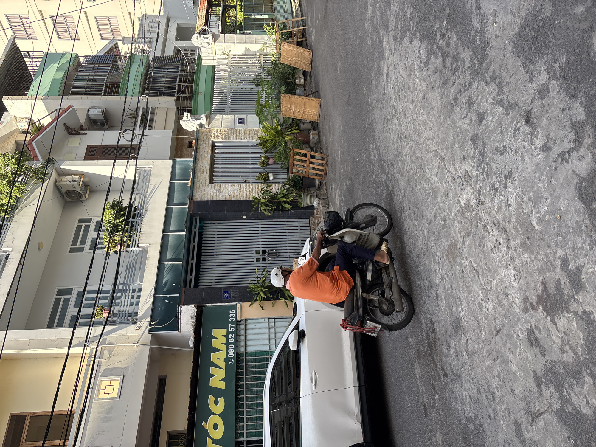 The image depicts a street scene in Nha Trang, Vietnam, with a focus on a man riding a motorcycle. The man, wearing an orange t-shirt, dark pants, and a white helmet, is the central subject, captured in motion. He is riding a dark-colored motorcycle with a white seat cover and a red rope tied to its rear. To the left of the motorcycle is a white car parked parallel to the street.
The background features a row of buildings, indicative of a residential or commercial area in the city. One building has a green awning with yellow lettering that reads "TÓC NAM" and a phone number "090 52 57 336". Above the awning, windows and balconies are visible on the multi-story structures. Power lines crisscross the scene above the buildings.
The street itself is paved with asphalt and shows signs of wear and tear, with patches of lighter-colored concrete or repairs. Several wooden pallets and other miscellaneous wooden items are placed on the sidewalk near the entrance to one of the buildings, which has a gray metal gate. Lush green plants are visible in pots and in the ground along the building's facade. The lighting suggests it is daytime, with sunlight casting shadows.
