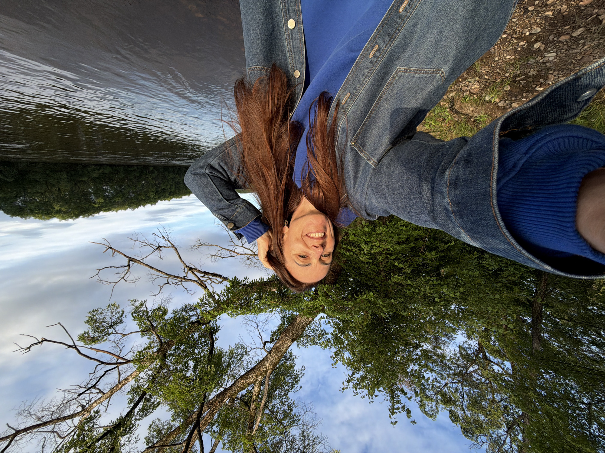 A mid-adult female is captured in a wide-angle selfie, smiling directly at the camera while outdoors in the city of Birobidzhan, Russia. She possesses long, reddish-brown hair and is dressed in a royal blue hooded sweatshirt underneath a denim jacket. Her left arm, partially visible in the lower-left corner, is extended to hold the camera, while her right hand is positioned near her hair on the right side of her head. The background features a wide, dark body of water, indicative of a river, occupying the right portion of the frame, with a dense line of green trees on the far bank. The immediate ground in the lower-left is composed of earth and small stones with scattered green vegetation. Overhead and to the left, a dense canopy of green trees is visible. Above the female and to the right, a large tree displays both green foliage and bare branches against a sky with scattered white and grey clouds. The scene is illuminated by natural daylight.