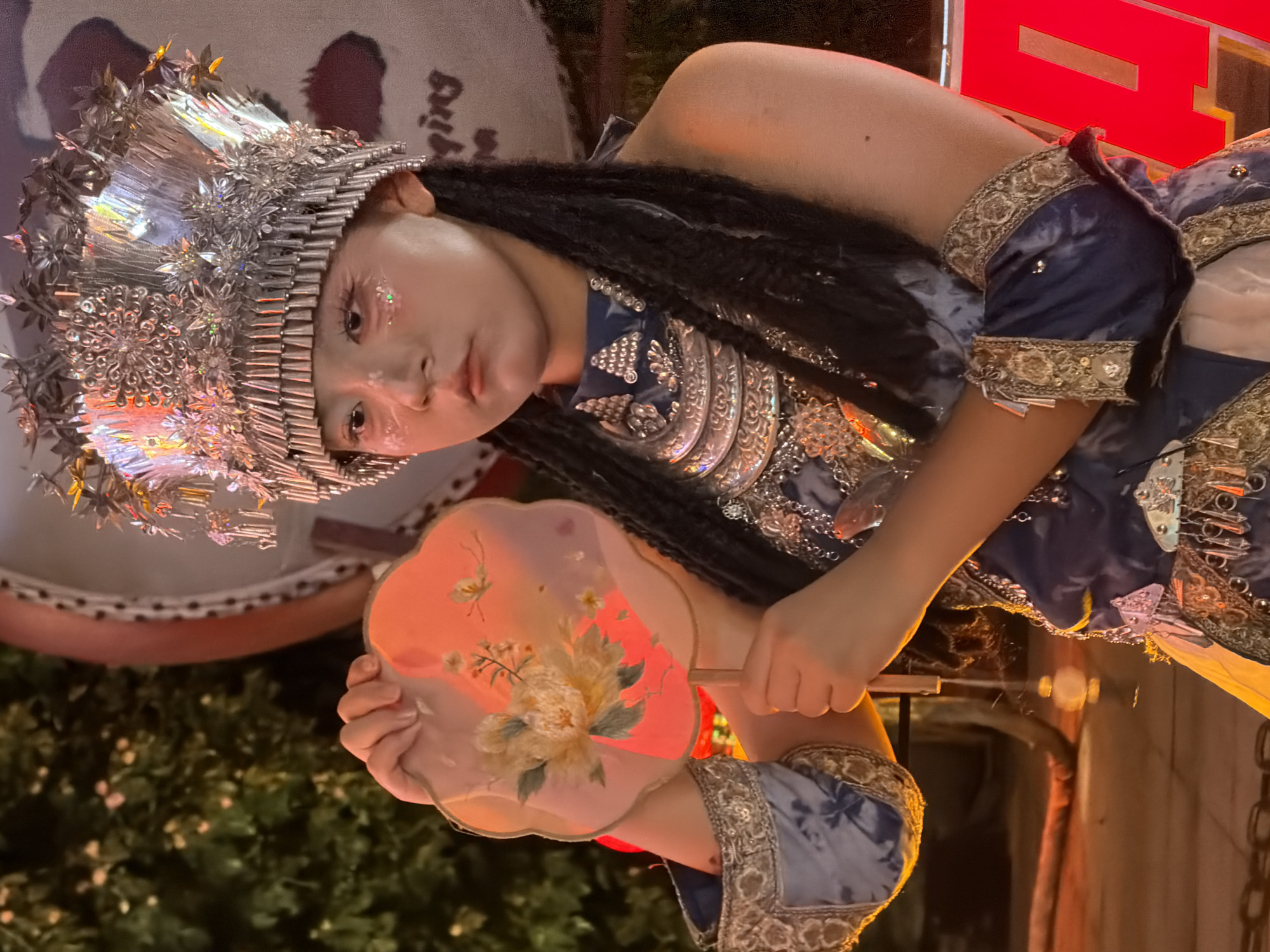 A young person, adorned in an ornate silver headdress and a blue, gold-trimmed costume, holds a delicate fan painted with flowers. Their face, with detailed eye makeup including glittery accents, is turned towards the viewer, conveying a calm and perhaps slightly solemn expression. The headdress, a complex structure of metal pieces and embellishments, shines brightly, reflecting the surrounding light. The costume features intricate silver detailing on the bodice and sleeves, adding to the regal or ceremonial appearance.
The fan, held in the person's hands, is translucent and illuminated from behind, highlighting the floral artwork. The background is blurred but suggests an outdoor setting, possibly at dusk or night, with hints of greenery and illuminated structures. The lighting is warm and atmospheric, casting glows on the subject and their attire. The overall impression is one of a cultural performance or a themed event, possibly taking place in Yuzhong District, China, given the provided location context. There is no visible text in the image.